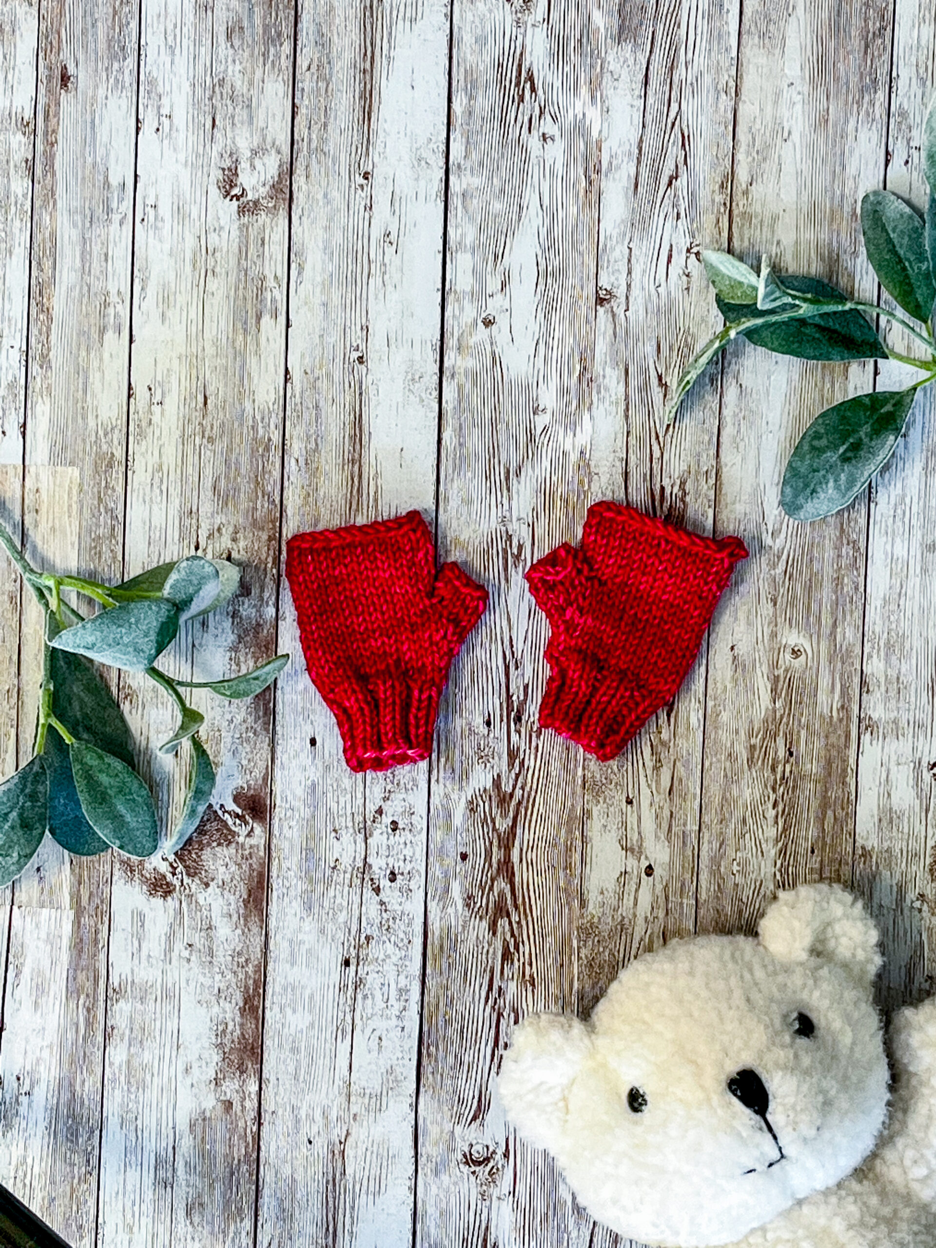 A pair of fuchsia, hand dyed merino fingerless mitts rests on a wood background with sage leaves and a white teddy bear face around it
