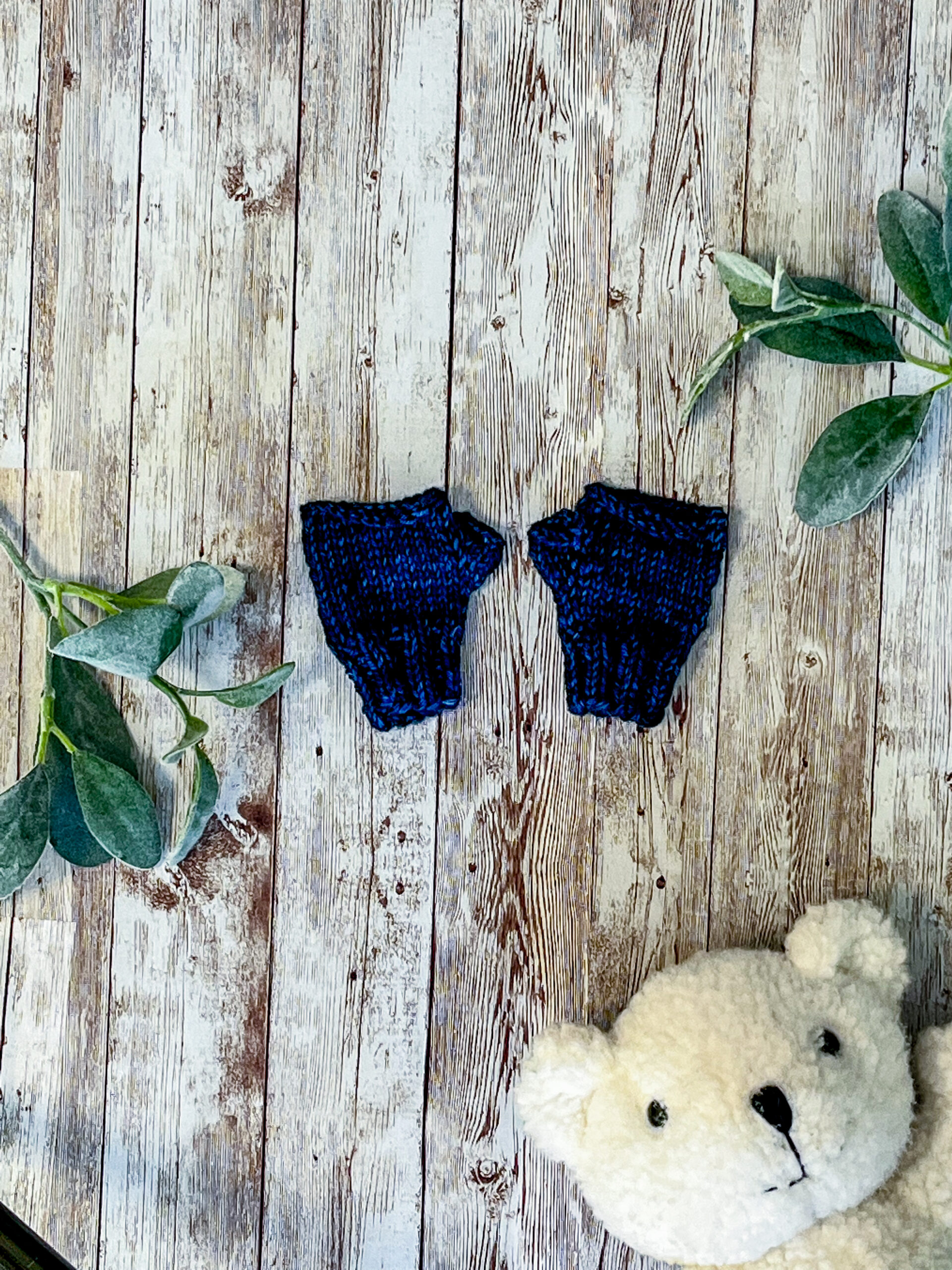 A pair of blue, hand dyed merino fingerless mitts rests on a wood background with sage leaves and a white teddy bear face around it