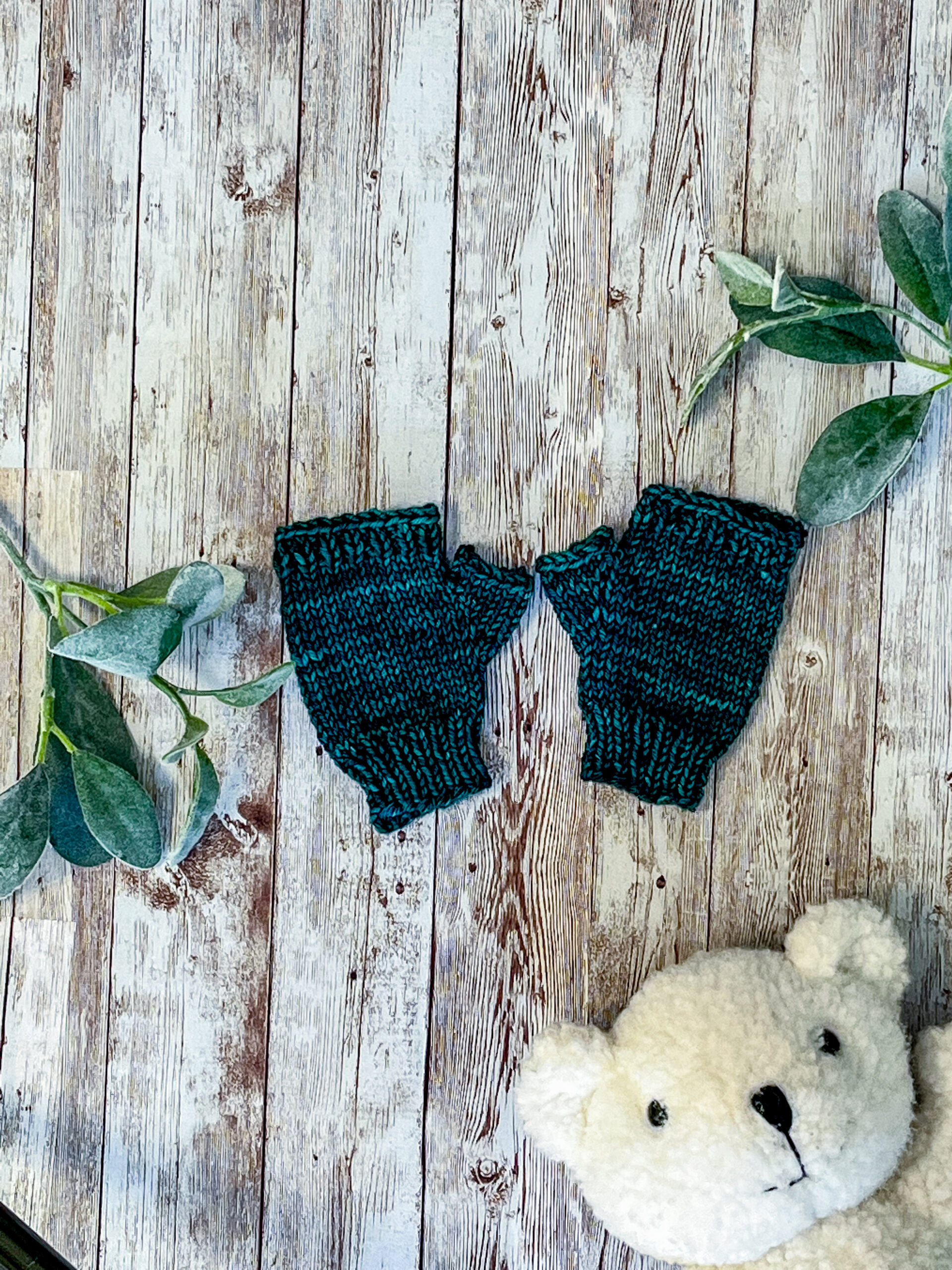 A pair of green, hand dyed merino fingerless mitts rests on a wood background with sage leaves and a white teddy bear face around it