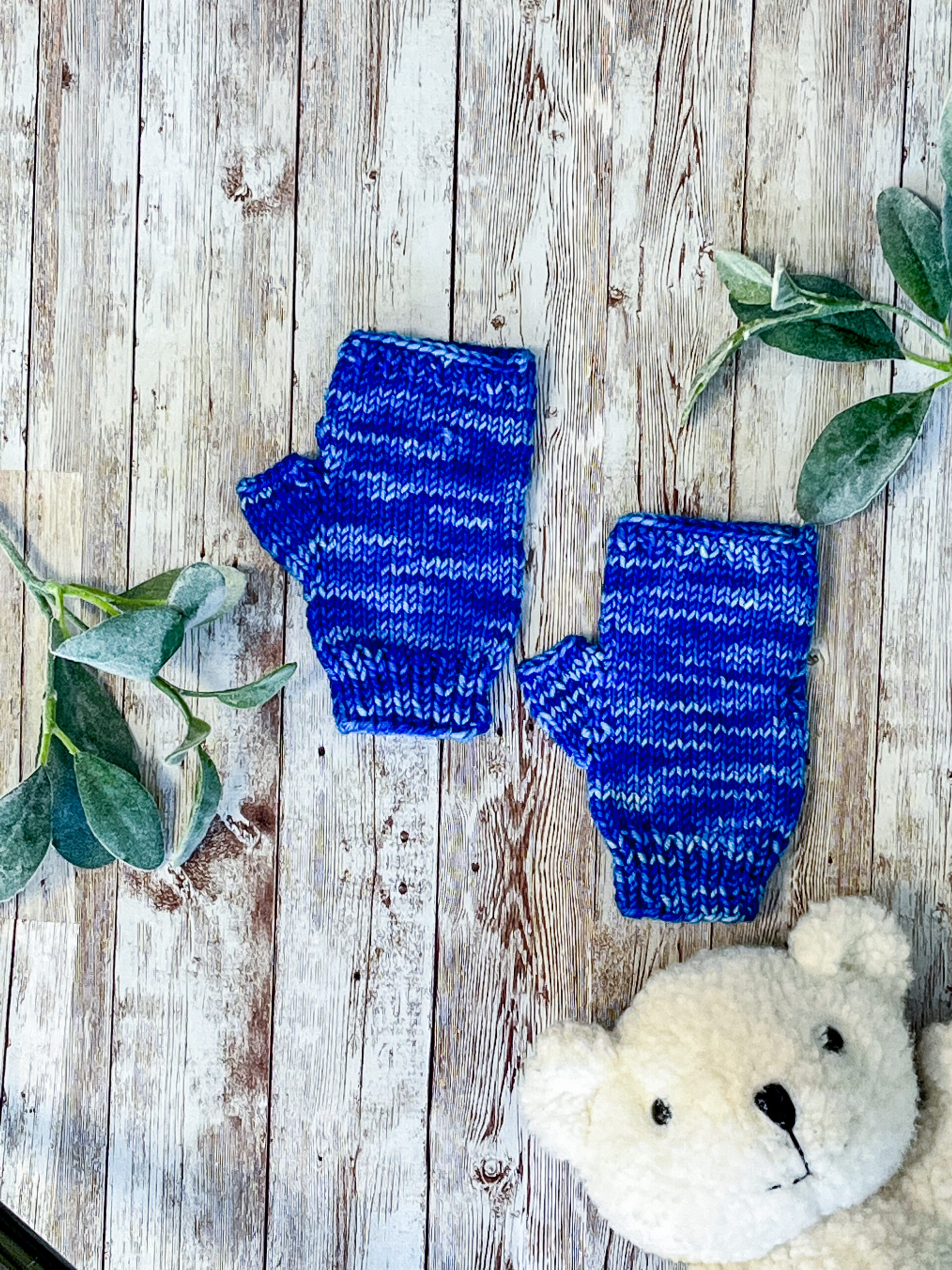 A pair of blue, hand dyed merino fingerless mitts rests on a wood background with sage leaves and a white teddy bear face around it