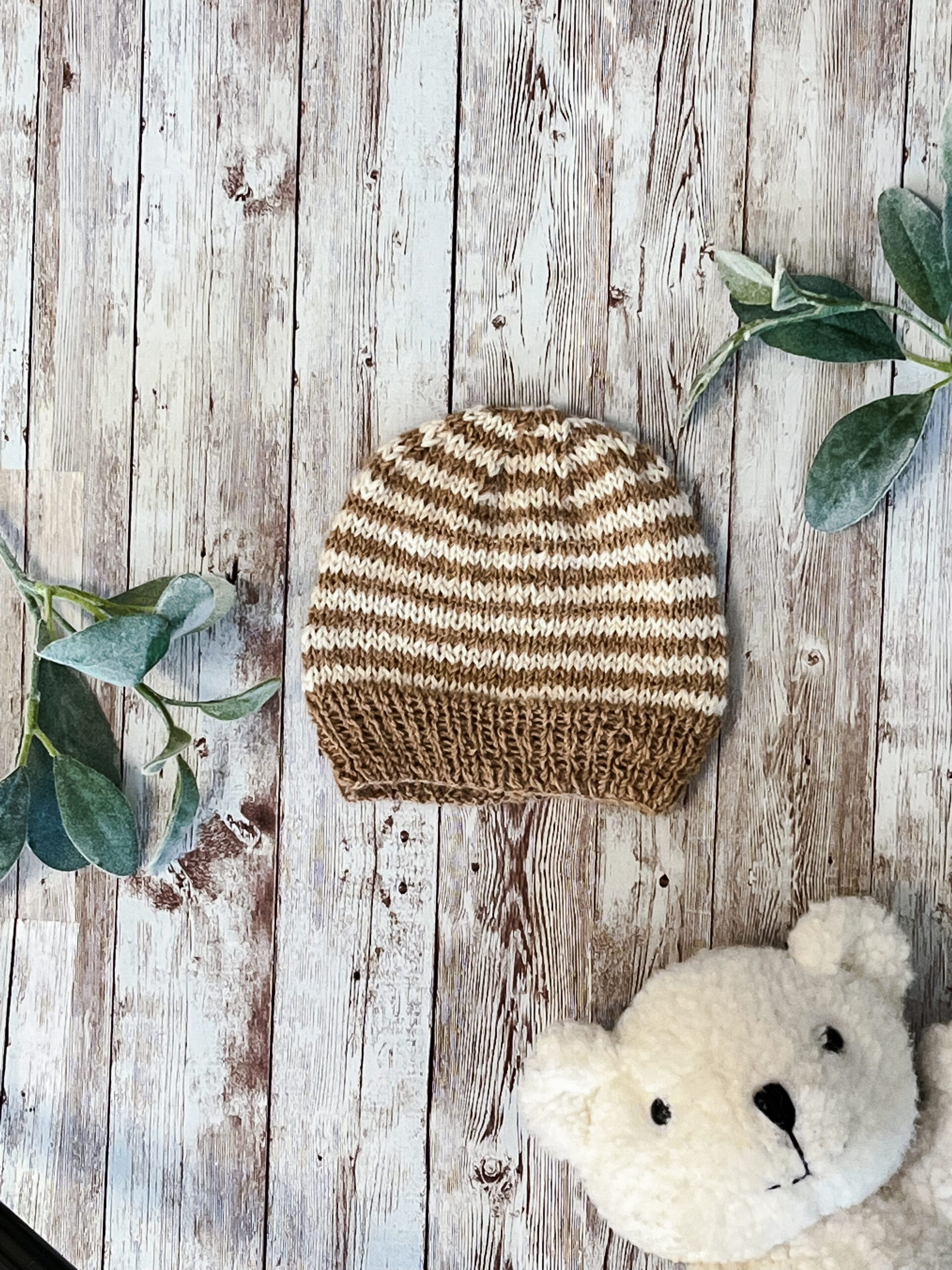 An alpaca hat with undyed tan and off-white strips is on a wood background with sage leaves and a white teddy bear