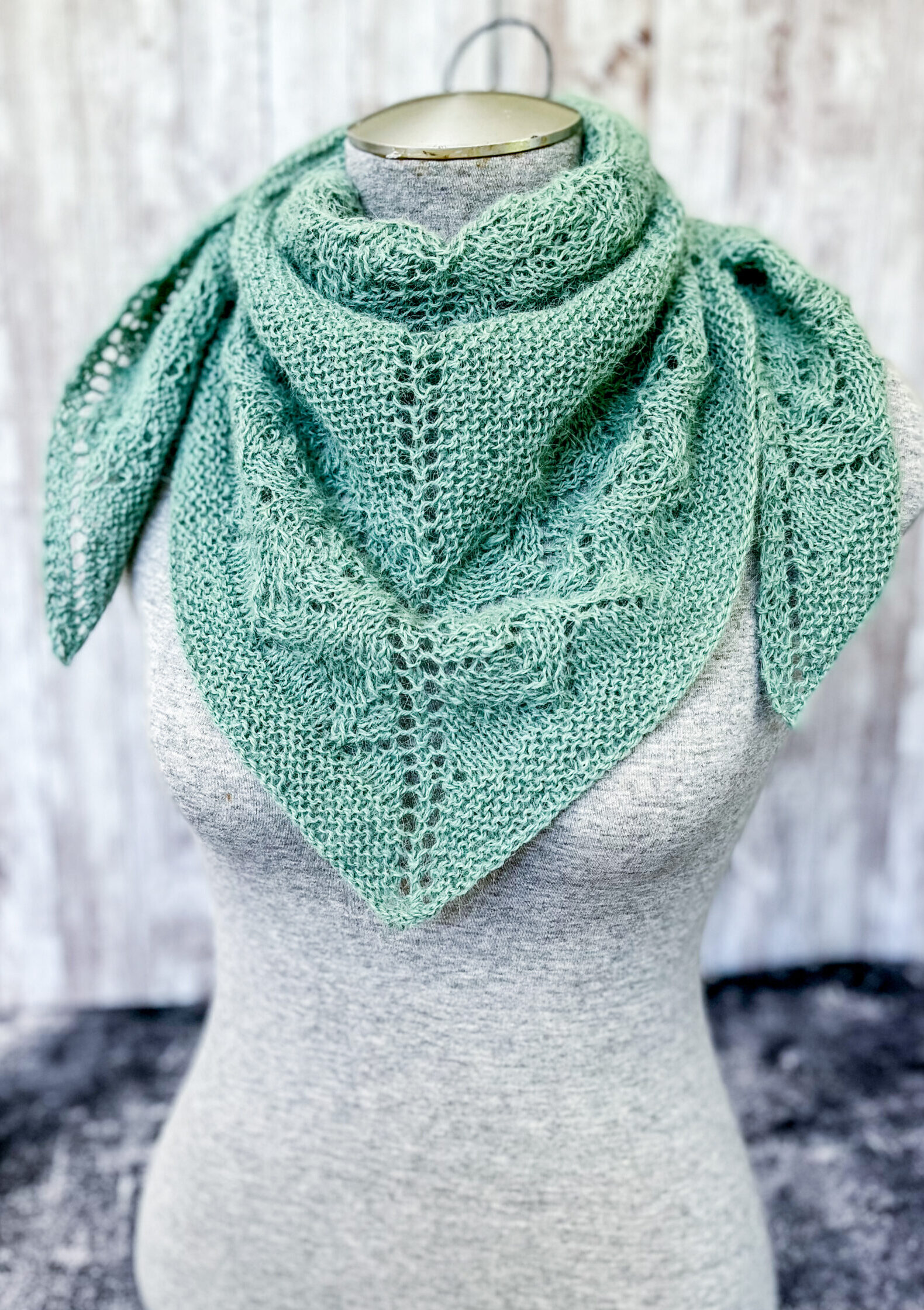 A light eucalyptus green shawl with lace sections alternating with solid sections is shown wrapped and gathered around the neck of a mannequin torso and pinned with a silver shawl pin