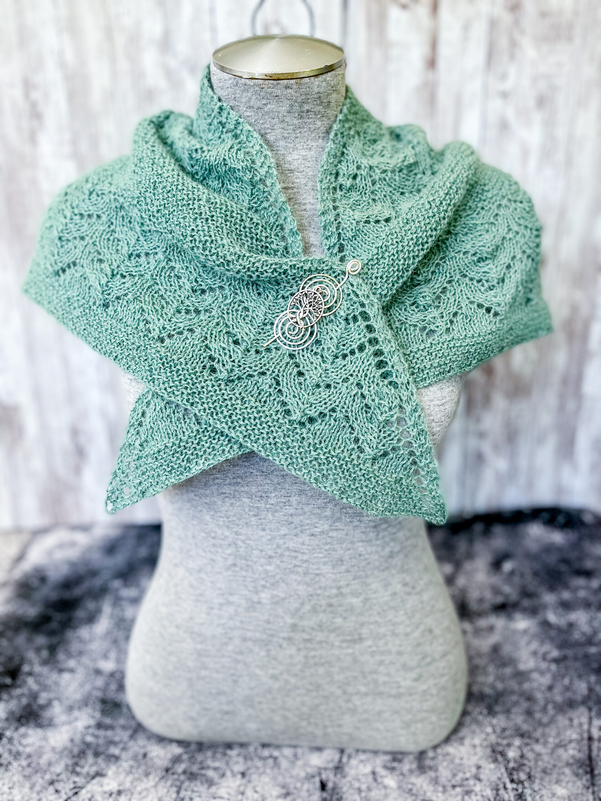 A light eucalyptus green shawl with lace sections alternating with solid sections is shown wrapped and crossing over around a mannequin torso and pinned with a silver shawl pin