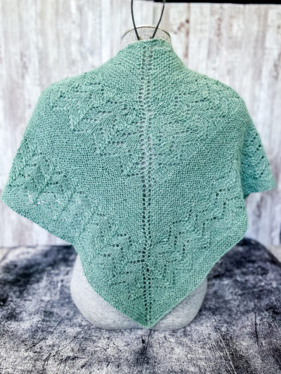 A light eucalyptus green shawl with lace sections alternating with solid sections is shown draped across the back of a mannequin torso.