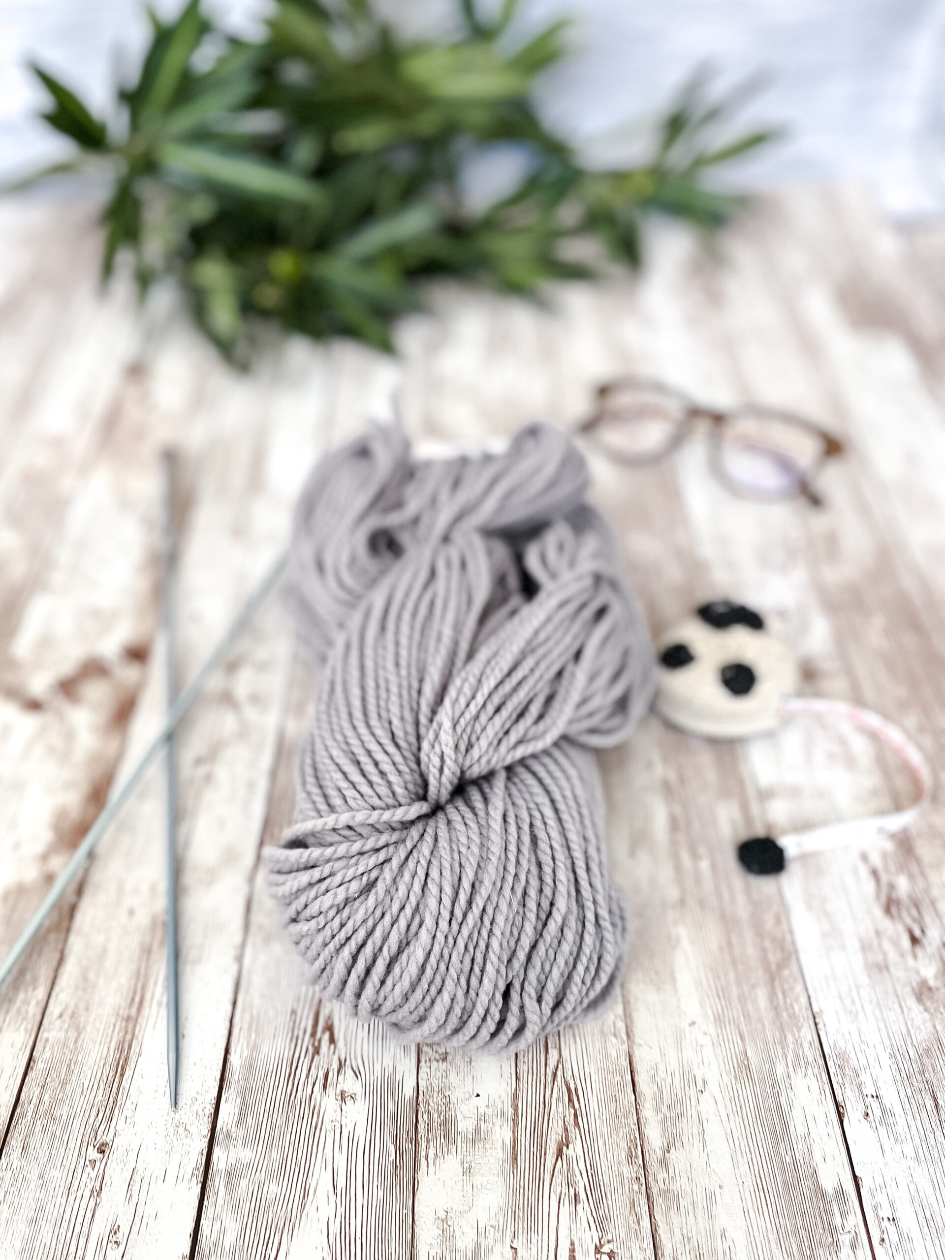 A hank of gray, Virginia farmed fine merino yarn rests on a wood plank, with knitting needles, a sheep measuring tape, a pair of reading glasses, and some greenery around it