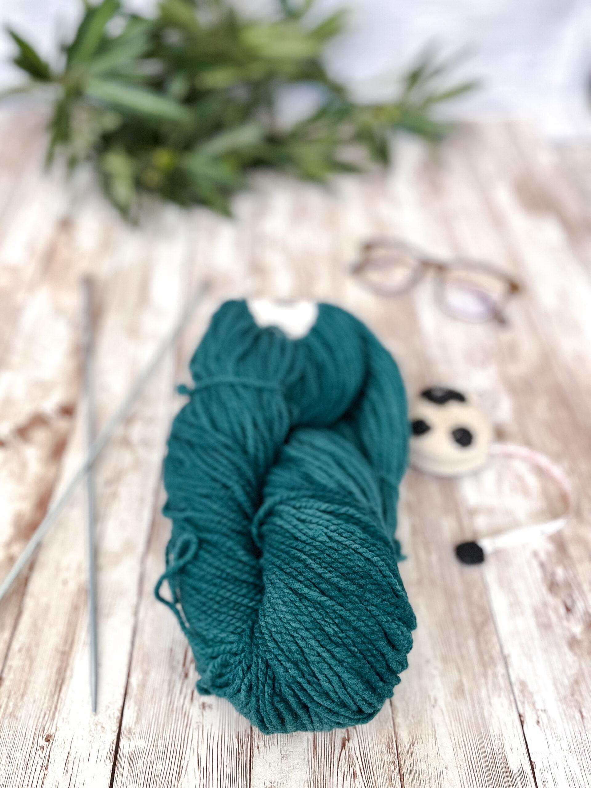 A hank of teal, Virginia farmed fine merino yarn rests on a wood plank, with knitting needles, a sheep measuring tape, a pair of reading glasses, and some greenery around it
