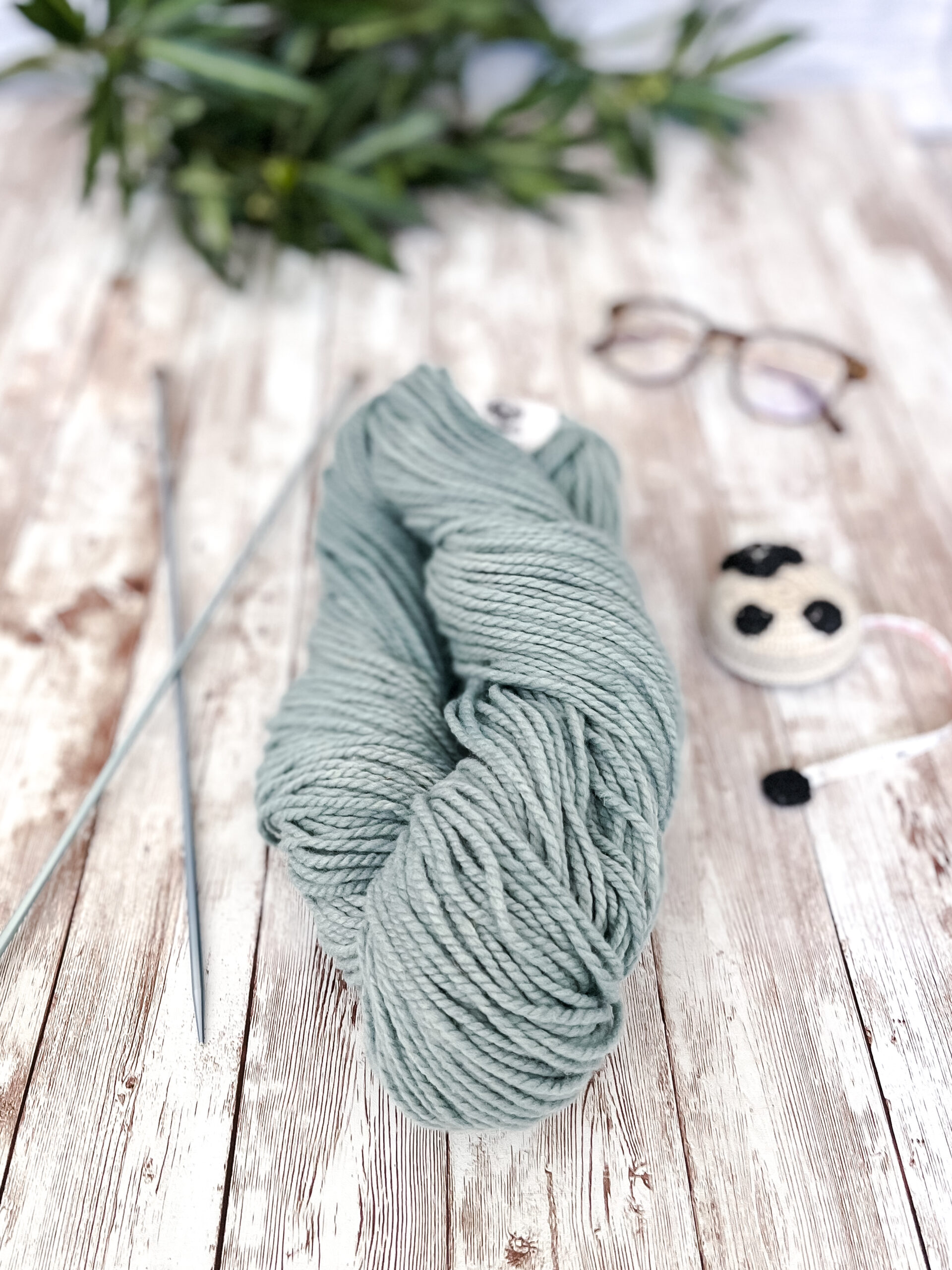A hank of sage green, Virginia farmed fine merino yarn rests on a wood plank, with knitting needles, a sheep measuring tape, a pair of reading glasses, and some greenery around it