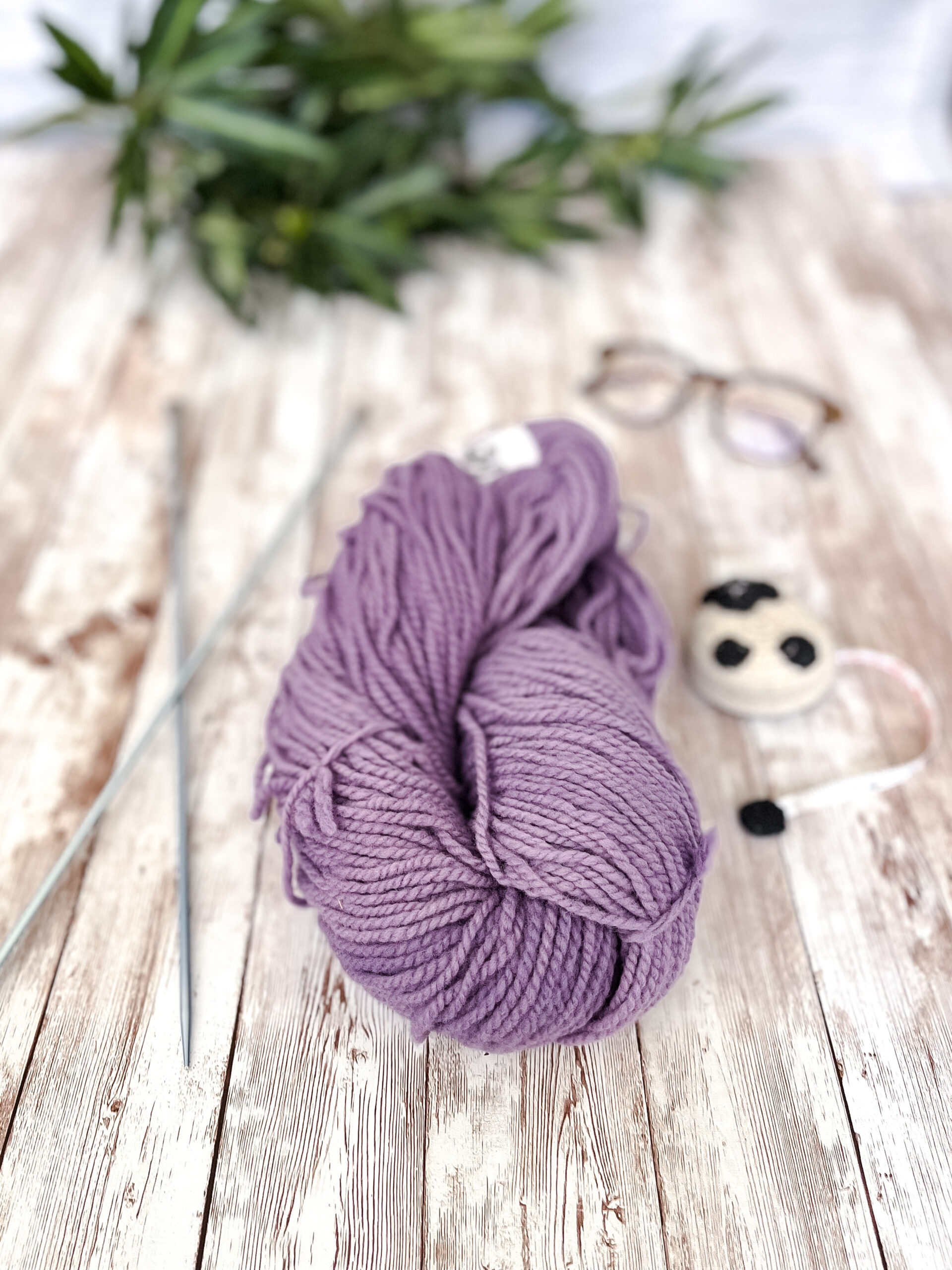 A hank of purple, Virginia farmed fine merino yarn rests on a wood plank, with knitting needles, a sheep measuring tape, a pair of reading glasses, and some greenery around it