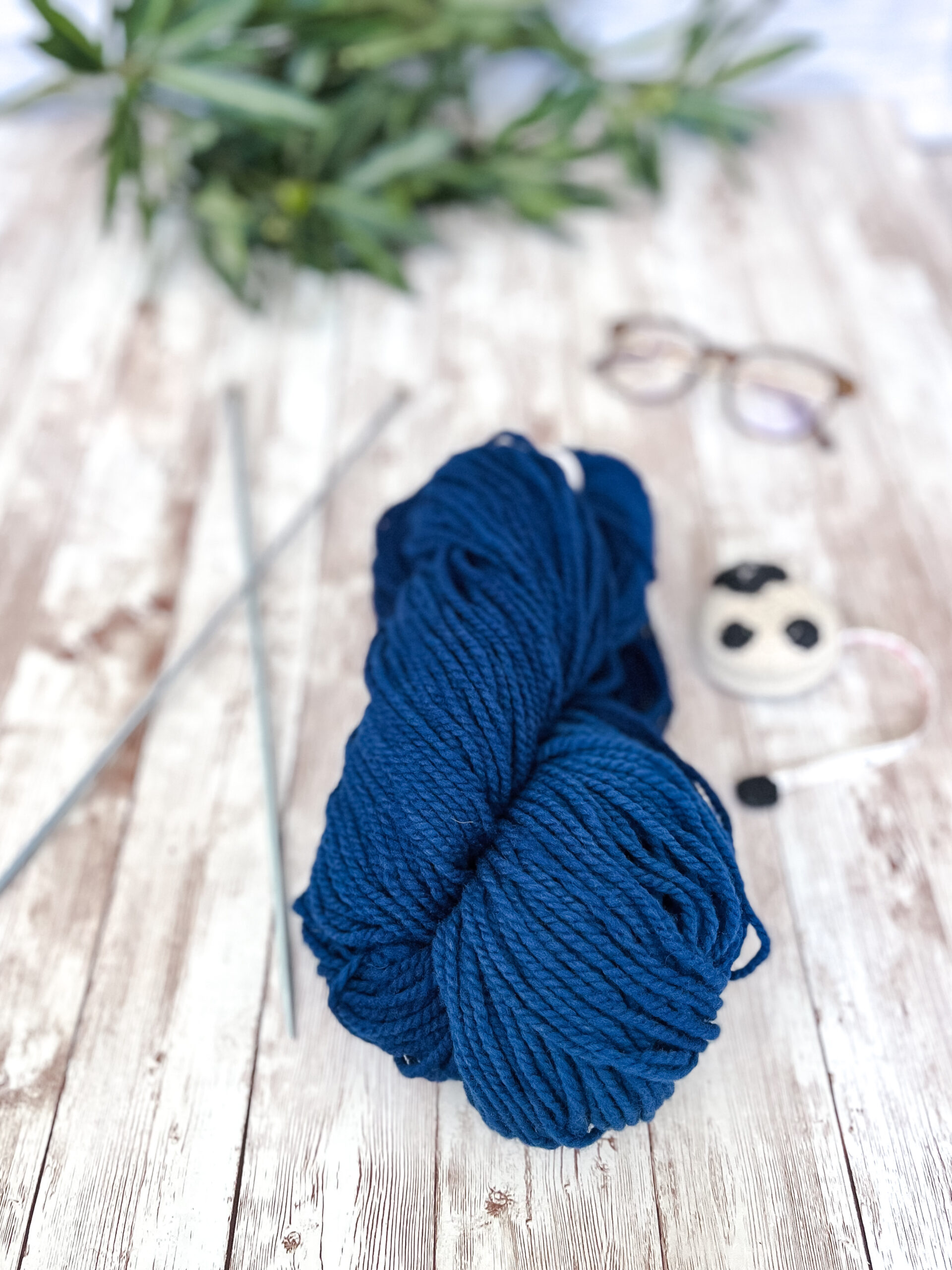 A hank of blue, Virginia farmed fine merino yarn rests on a wood plank, with knitting needles, a sheep measuring tape, a pair of reading glasses, and some greenery around it