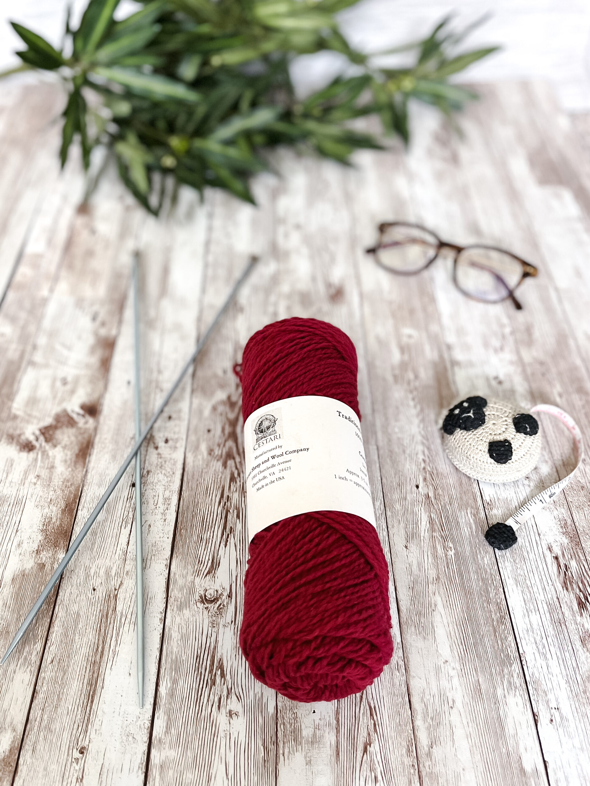 A red skein of yarn rests on a wood plank, with a pair of knitting needles, reading glasses, a lamb tape measure and greenery around it