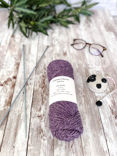 A light heathered purple skein of yarn rests on a wood plank, with a pair of knitting needles, reading glasses, a lamb tape measure and greenery around it