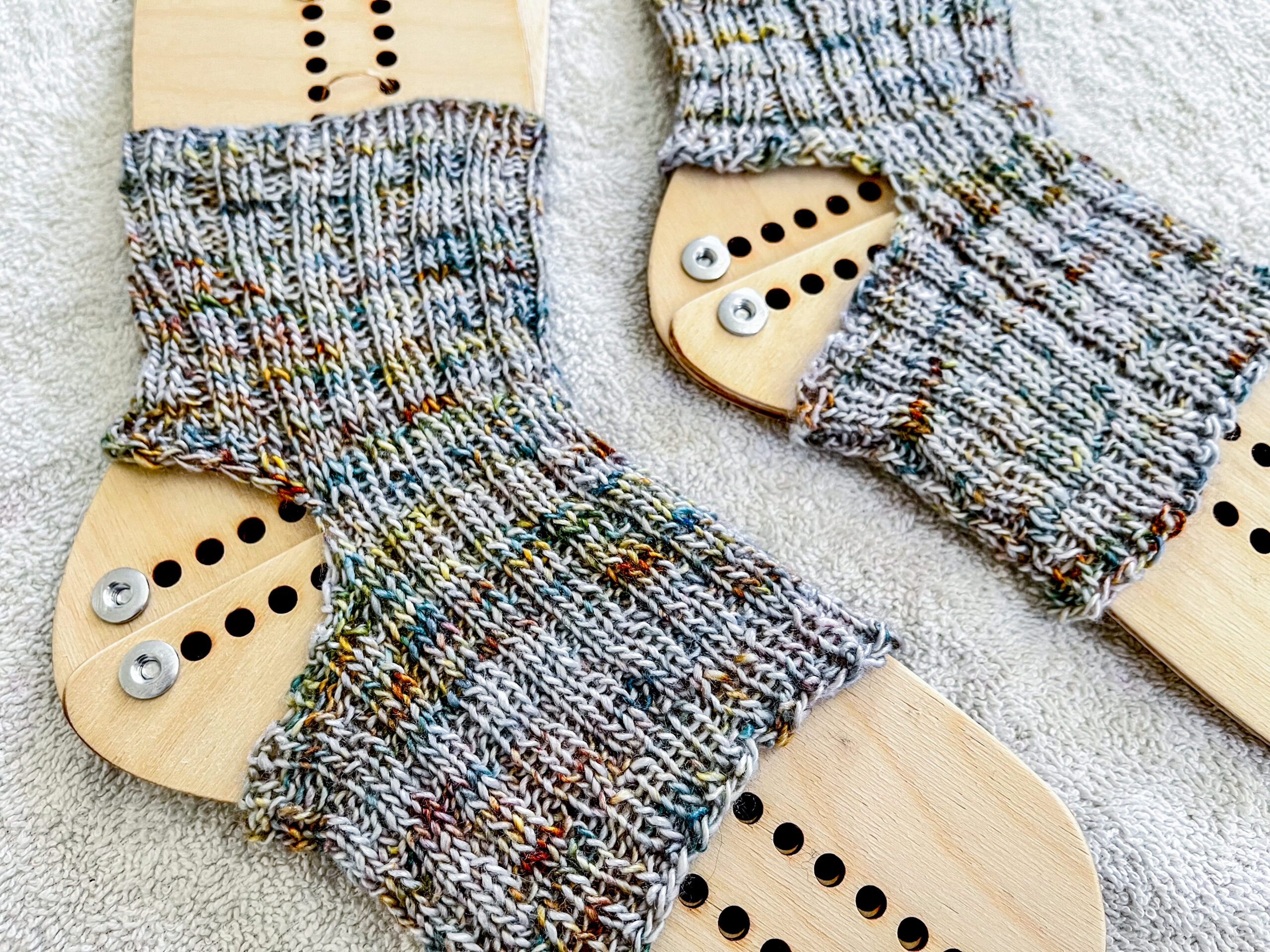 a pair of hand dyed merino/silk yoga socks are displayed on a set of wooden sock blockers. They are silver with flecks of teal, gold, and bronze speckled throughout the yarn.