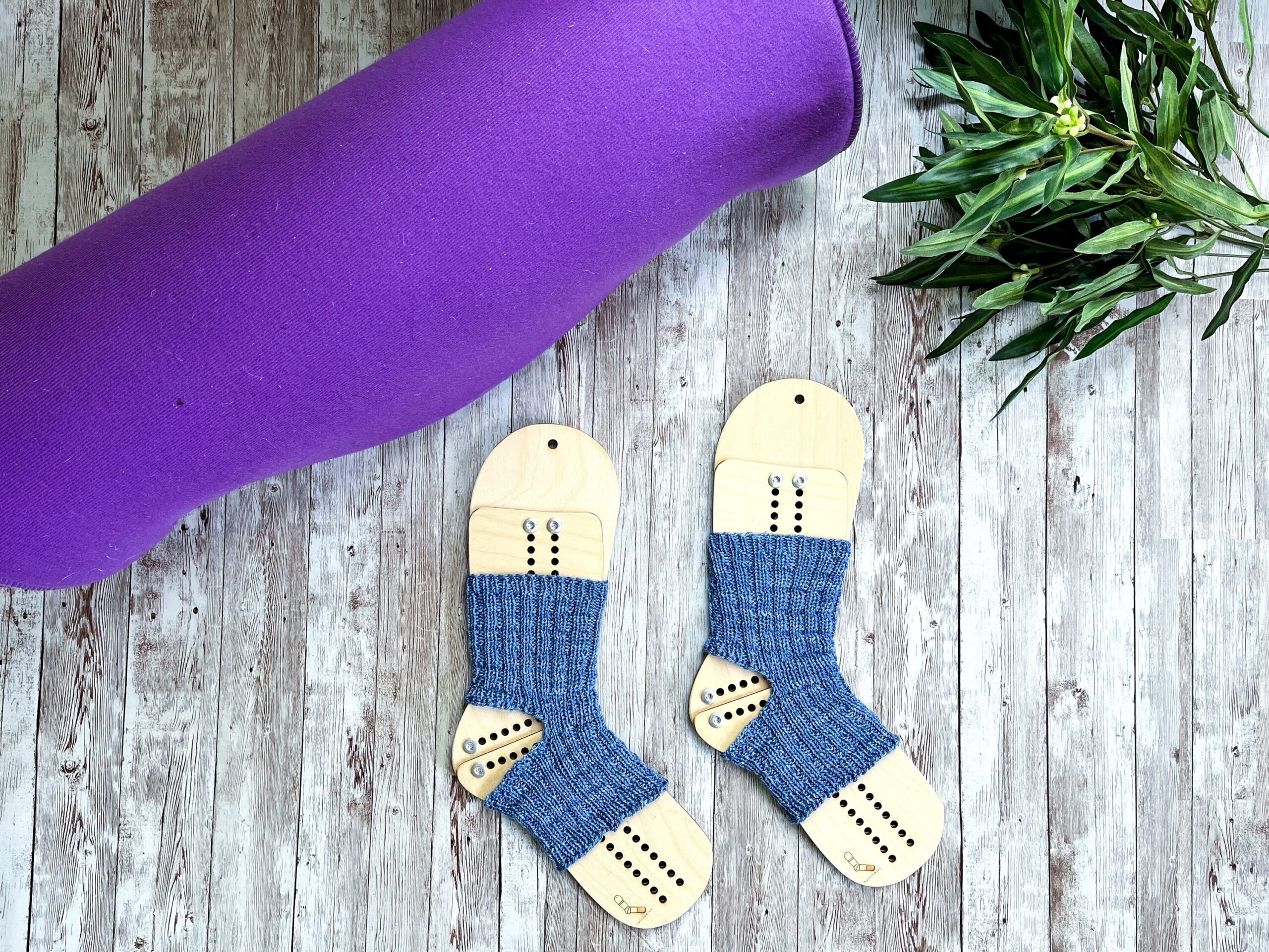 A pair of blue yoga socks on wooden sock blockers are surrounded by a rolled up yoga mat and greenery on a wood panel