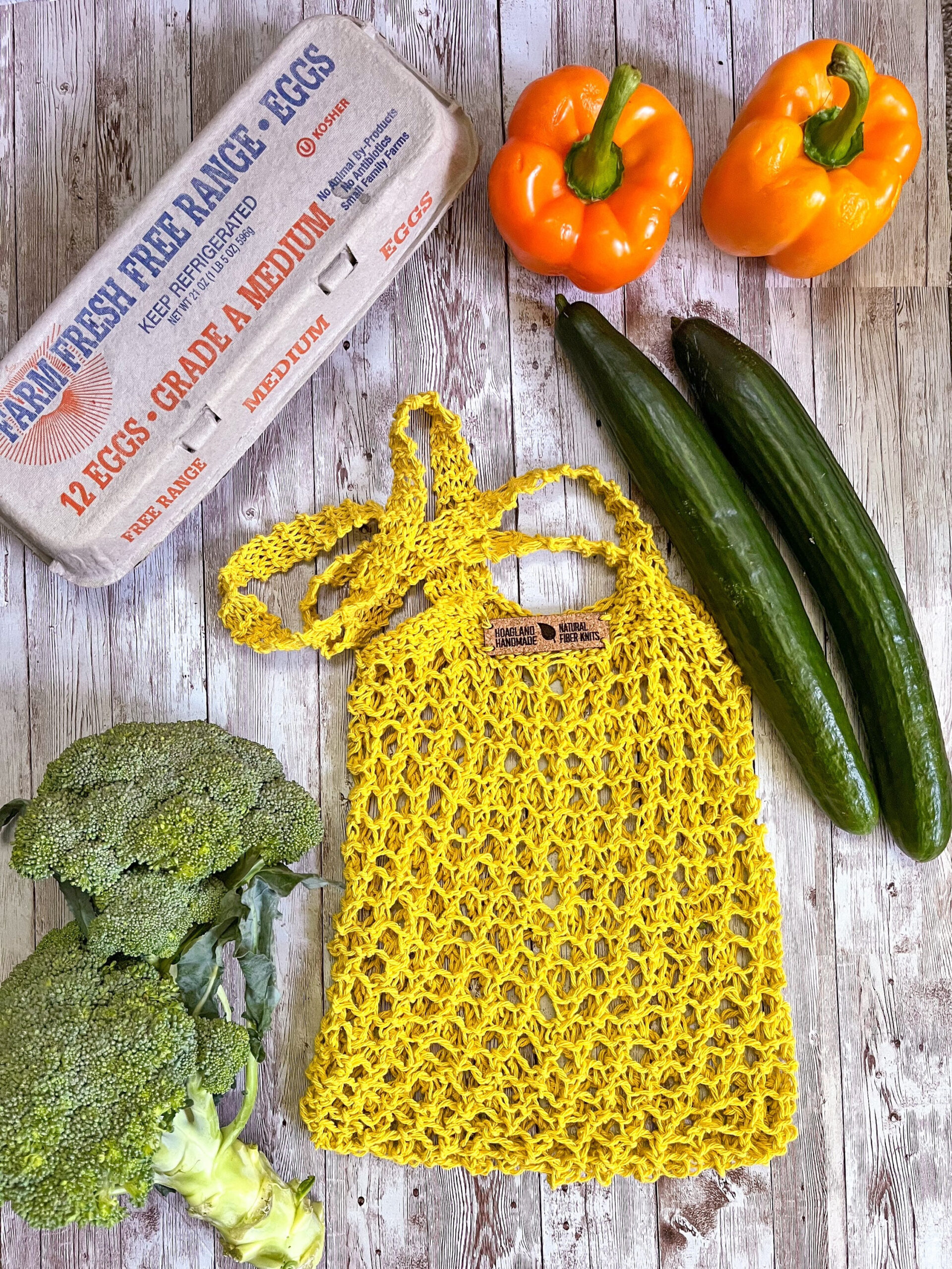 A yellow recycled cotton mesh tote bag is surrounded by a carton of farm eggs, orange bell peppers, cucumbers, and broccoli