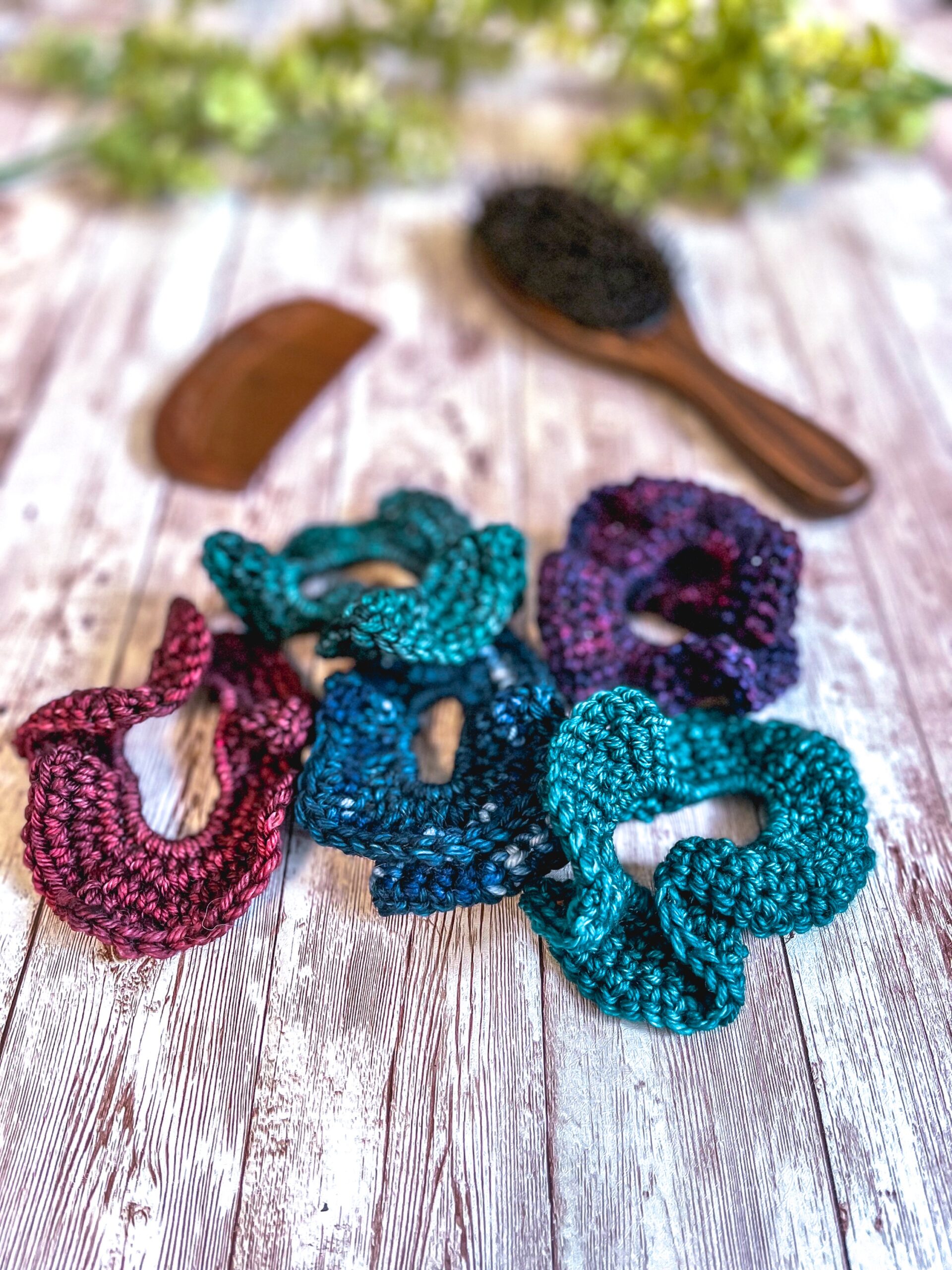 A set of 5 ruffled hair bands made with hand-dyed merino rests on a wood plank with a wood brush, wood comb, and greenery in the background. The earthy toned colors include a green, teal, purple, blue, and red