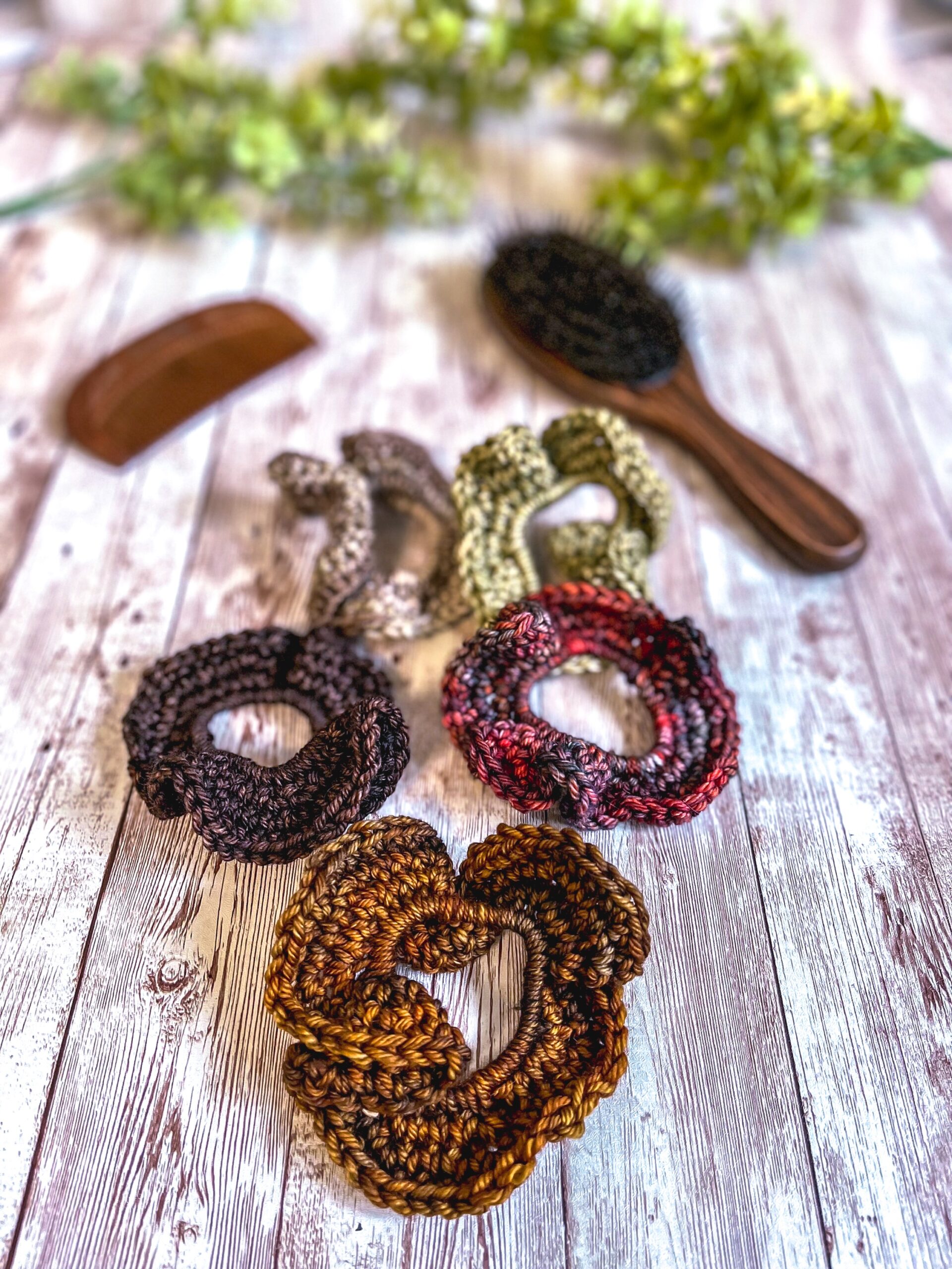 A set of 5 ruffled hair bands made with hand-dyed merino rests on a wood plank with a wood brush, wood comb, and greenery in the background. The earthy toned colors include a copper, brown, rustic green, brownish/gray, and a blend of oranges, reds, and browns.