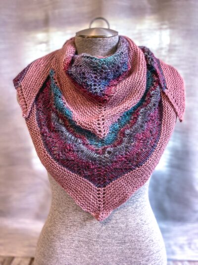A Blue Faced Leicester Wool shawl is draped around a gray mannequin torso. The shawl has solid blush pink sections alternated with a self-striping section with yarn in burgundy, purple, blue, teal and gray
