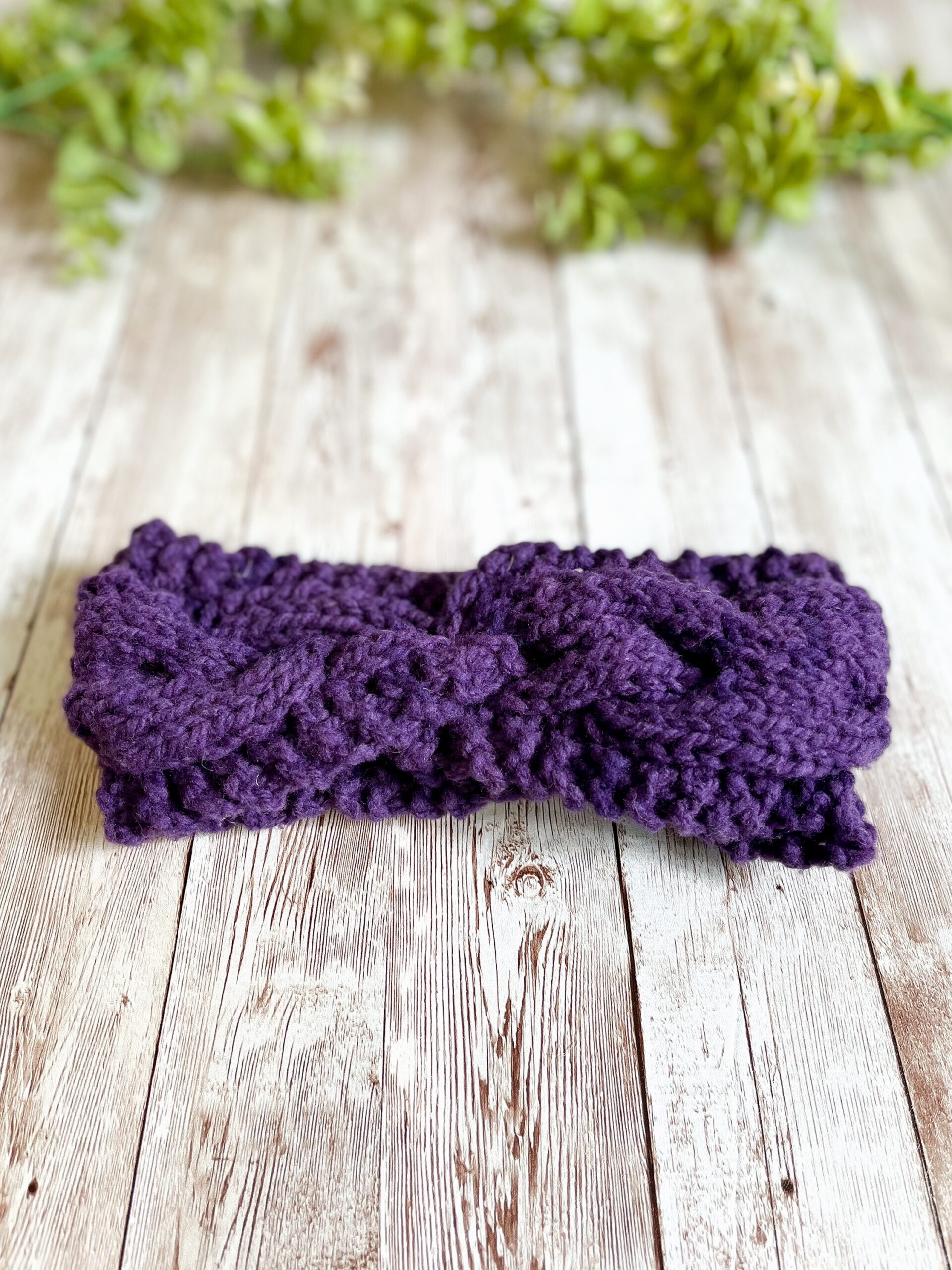 A purple, Virginia-raised merino ear warmer rests on a wood plank with greenery in the background
