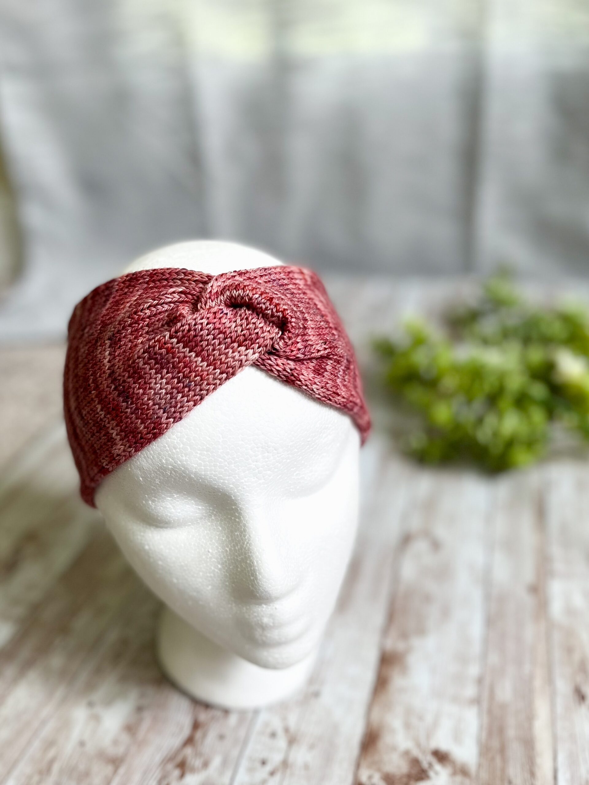 A mannequin head displays a reddish pink hand-dyed merino ear warmer