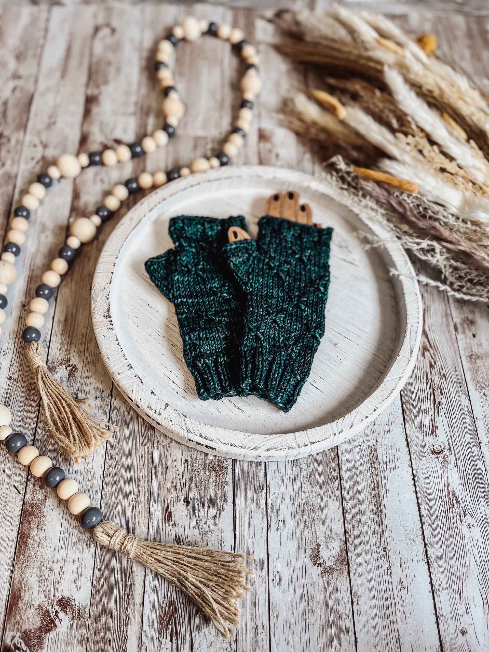 A pair of green, hand-dyed merino fingerless mitts rests on a white wood tray surrounded by fall pampas grass and wood beads