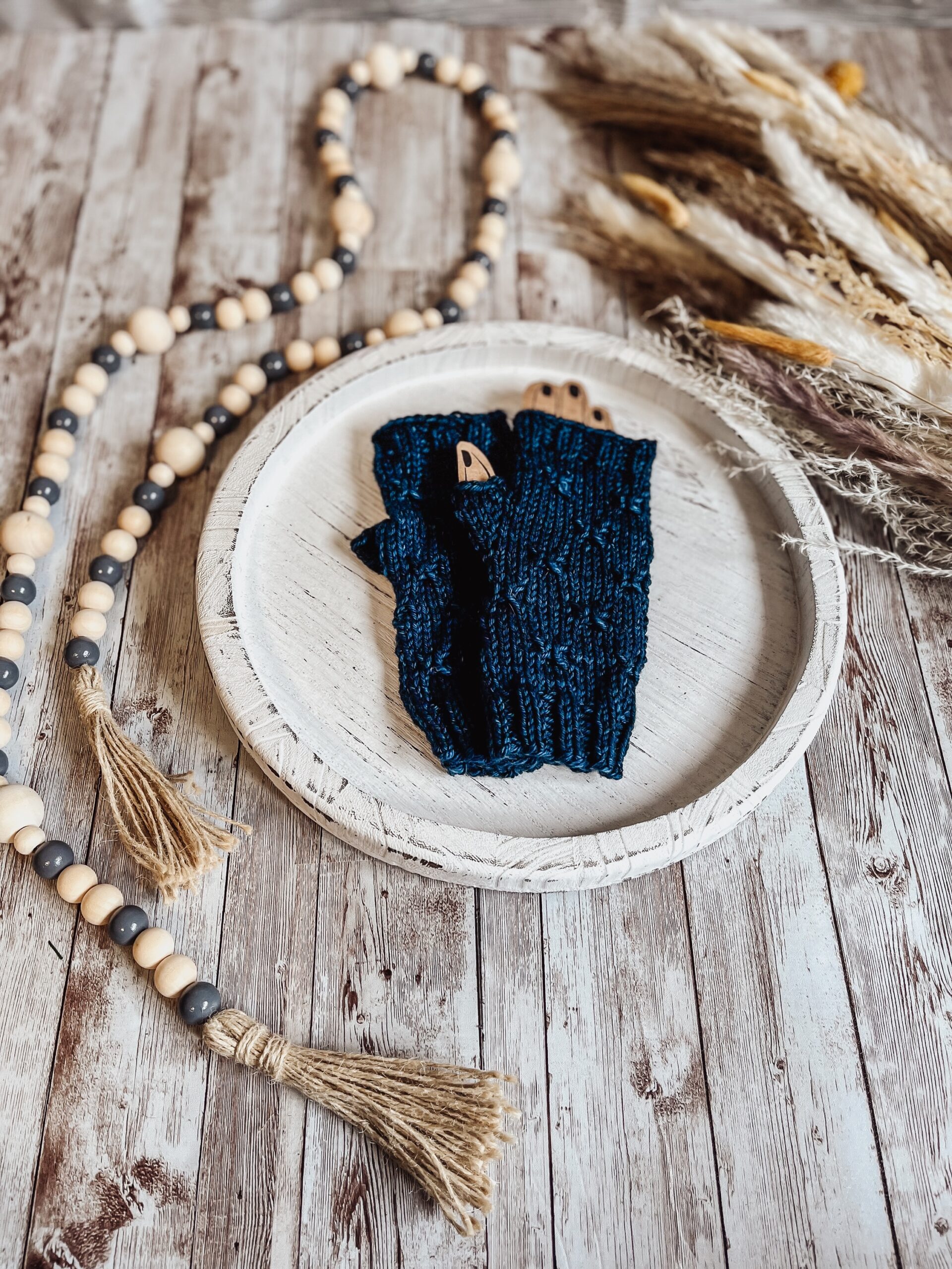A pair of blue, hand-dyed merino fingerless mitts rests on a white wood tray surrounded by fall pampas grass and wood beads