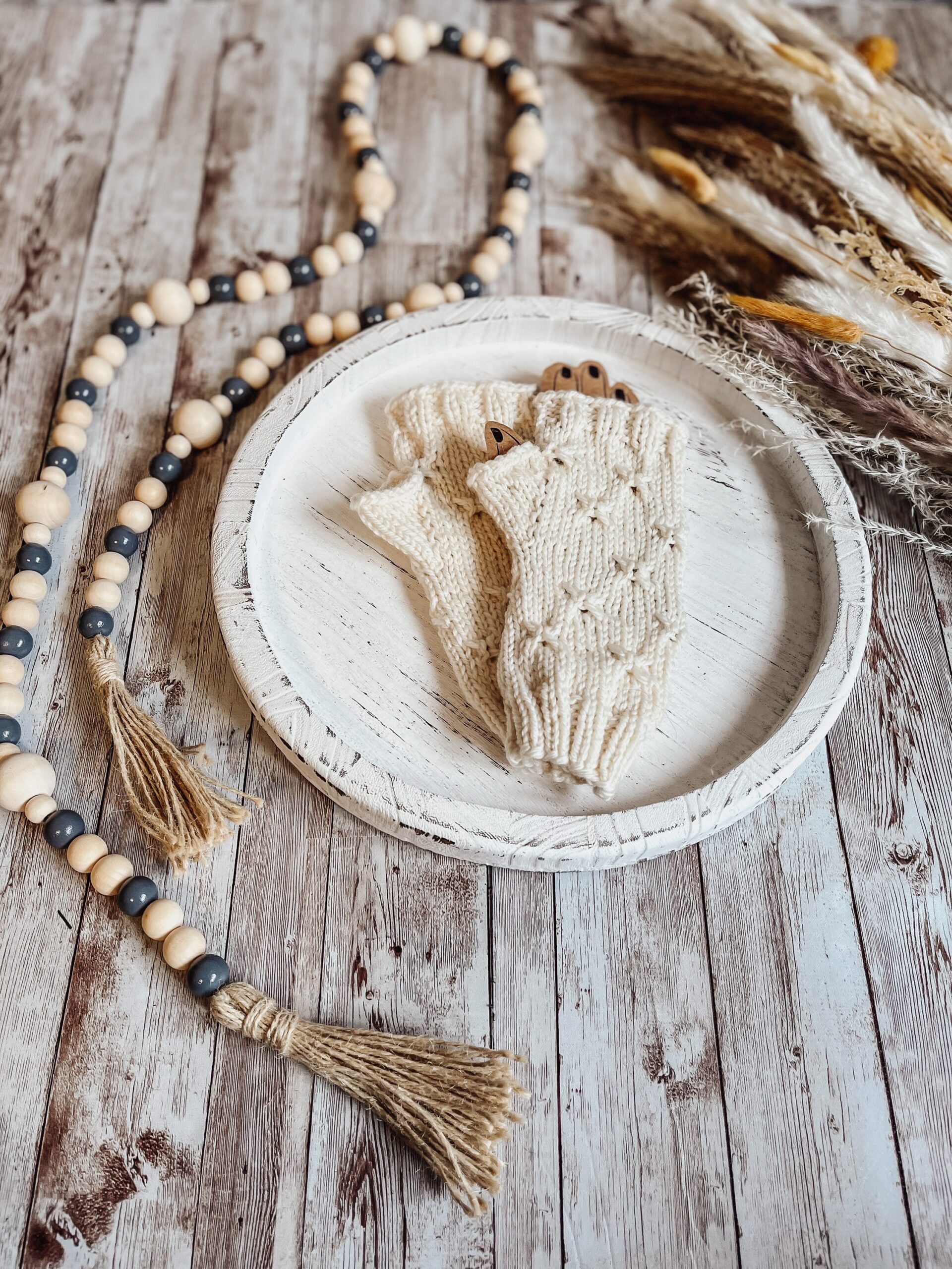 A pair of white, hand-dyed merino fingerless mitts rests on a white wood tray surrounded by fall pampas grass and wood beads