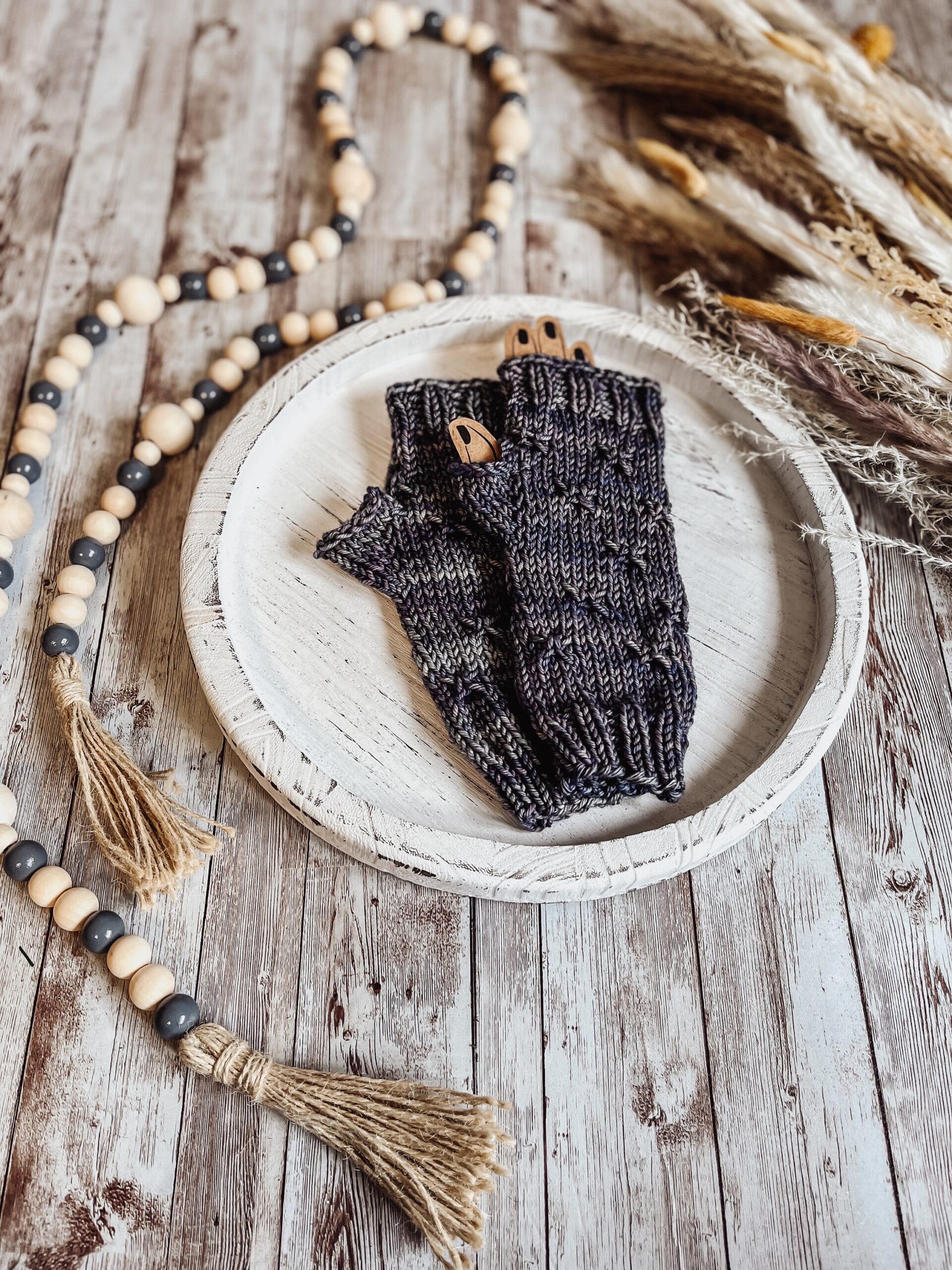 A pair of gray, hand-dyed merino fingerless mitts rests on a white wood tray surrounded by fall pampas grass and wood beads