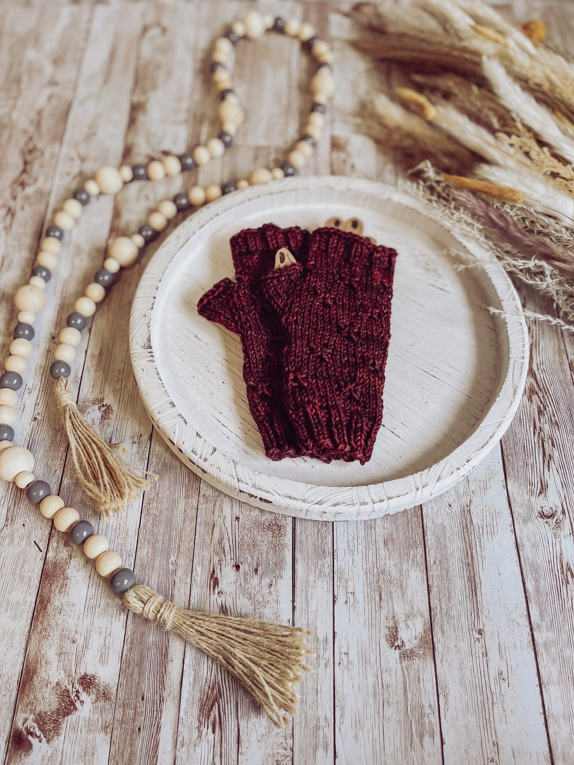 A pair of red, hand-dyed merino fingerless mitts rests on a white wood tray surrounded by fall pampas grass and wood beads T