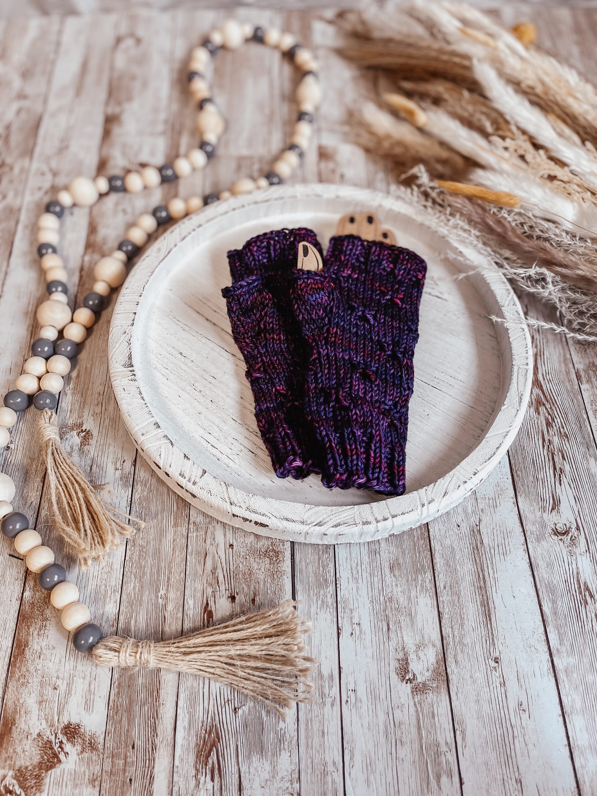 A pair of purple, hand-dyed merino fingerless mitts rests on a white wood tray surrounded by fall pampas grass and wood beads