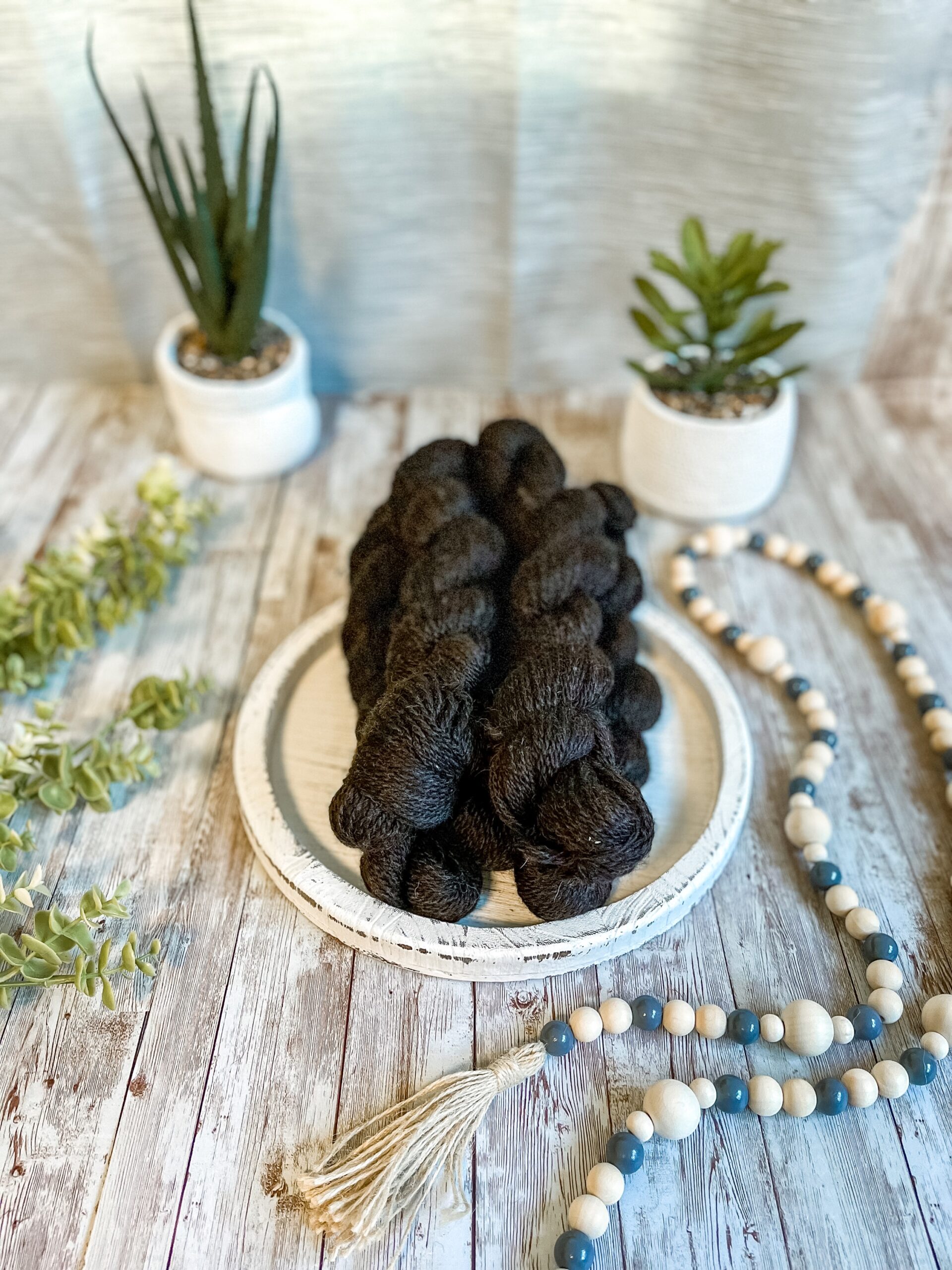Five skeins of natural black alpaca yarn rests on a white wood trivet. They are surrounded by beads to the right, greenery to the left and 2 succulent plants in the background.