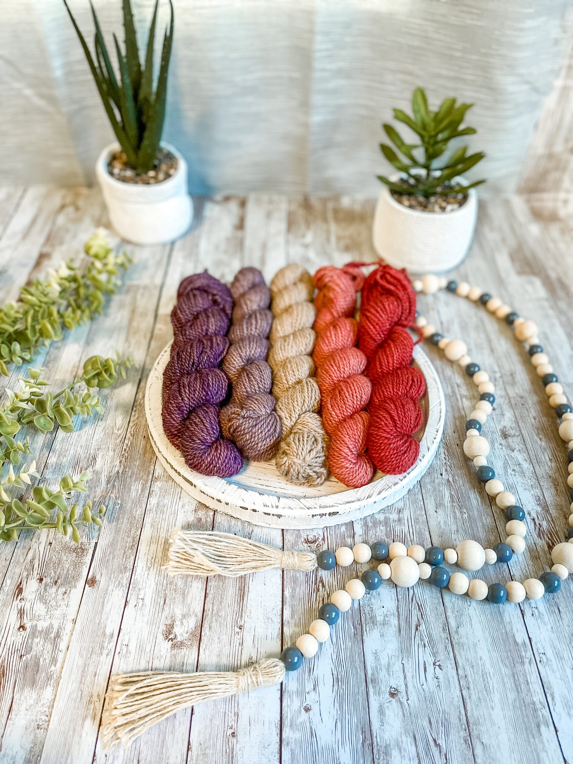 5 mini skeins of alpaca rest on a white wood trivet. They range in color from a dark purple, light purple, natural undyed fawn, light red, and dark red. They are surrounded by beads to the right, greenery to the left and 2 succulent plants in the background.