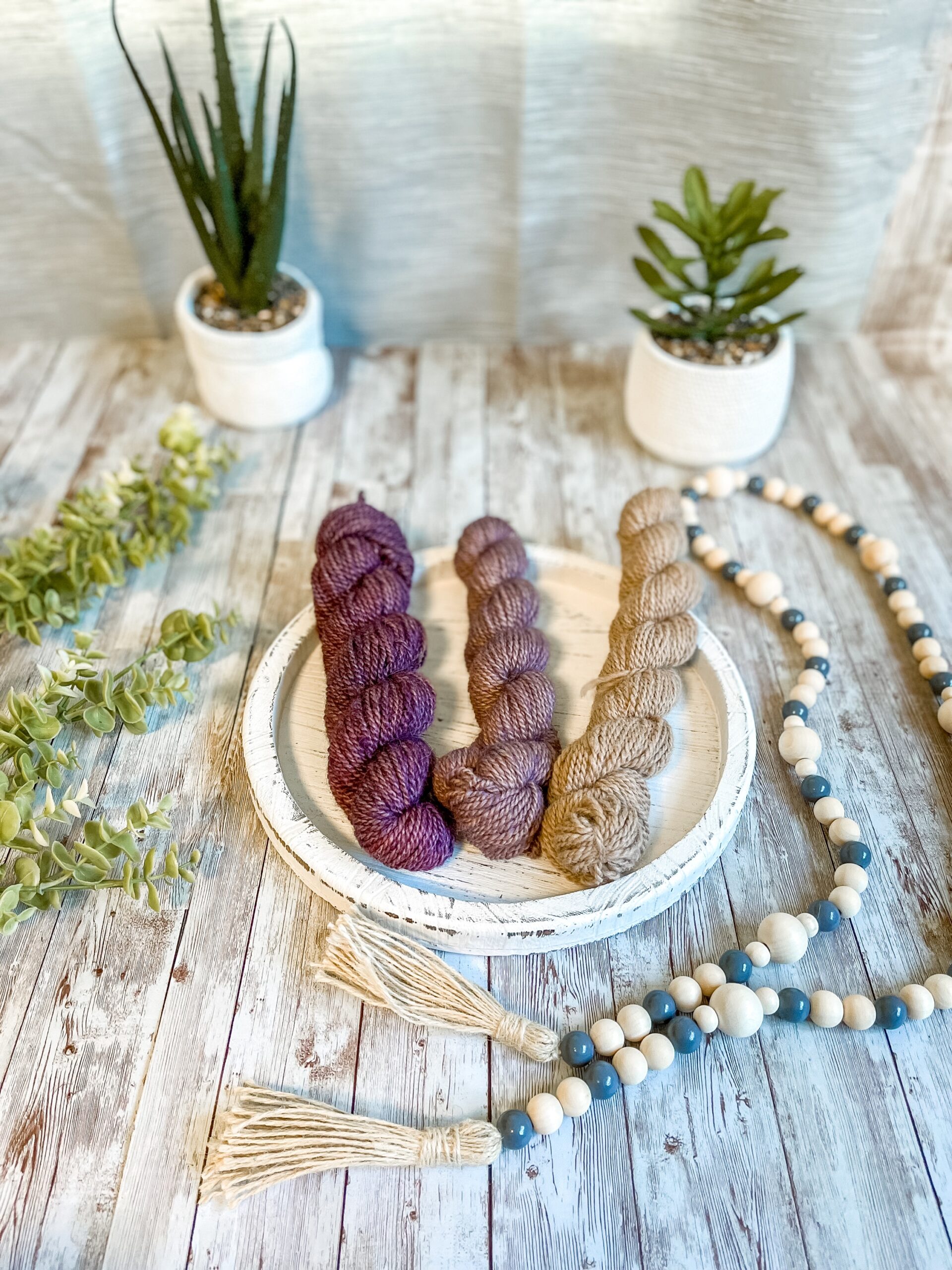 A set of 3 mini skeins in a purple fade from dark purple to natural fawn,rests on a white wood trivet. They are surrounded by beads to the right, greenery to the left and 2 succulent plants in the background.