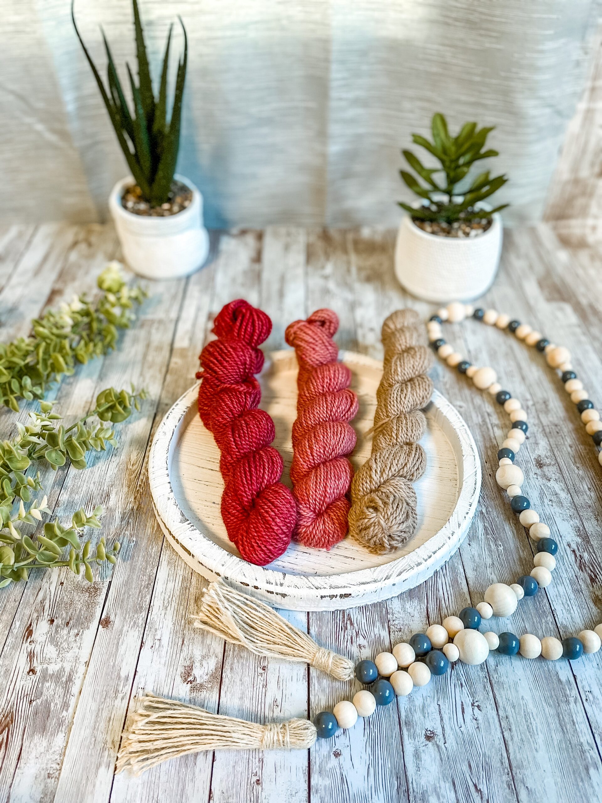 A set of 3 mini skeins in a red fade from dark purple to natural fawn,rests on a white wood trivet. They are surrounded by beads to the right, greenery to the left and 2 succulent plants in the background.