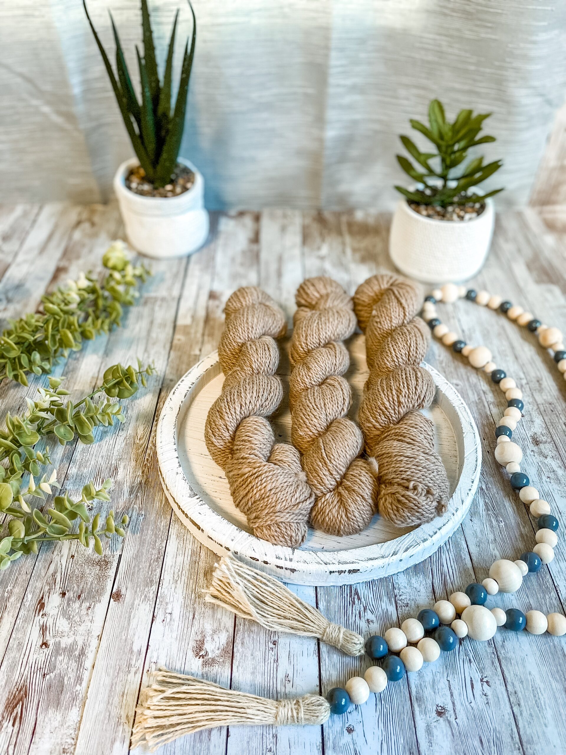 A set of 3 mini skeins of the natural fawn rests on a white wood trivet. They are surrounded by beads to the right, greenery to the left and 2 succulent plants in the background.
