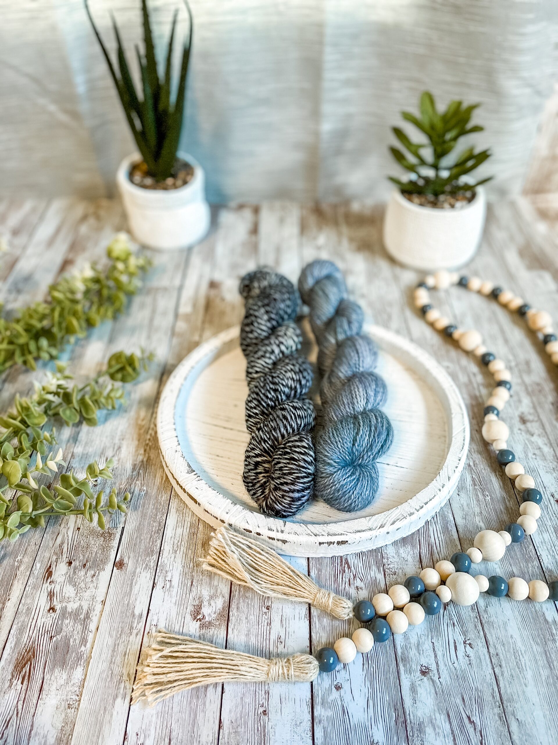 1 skein of hand-dyed, blue yarn and 1 twisted blue & black skein rest on a white wooden trivet. A strand of beads is draped to the right, some greenery is on the left, and 2 succulent plants are in the background.