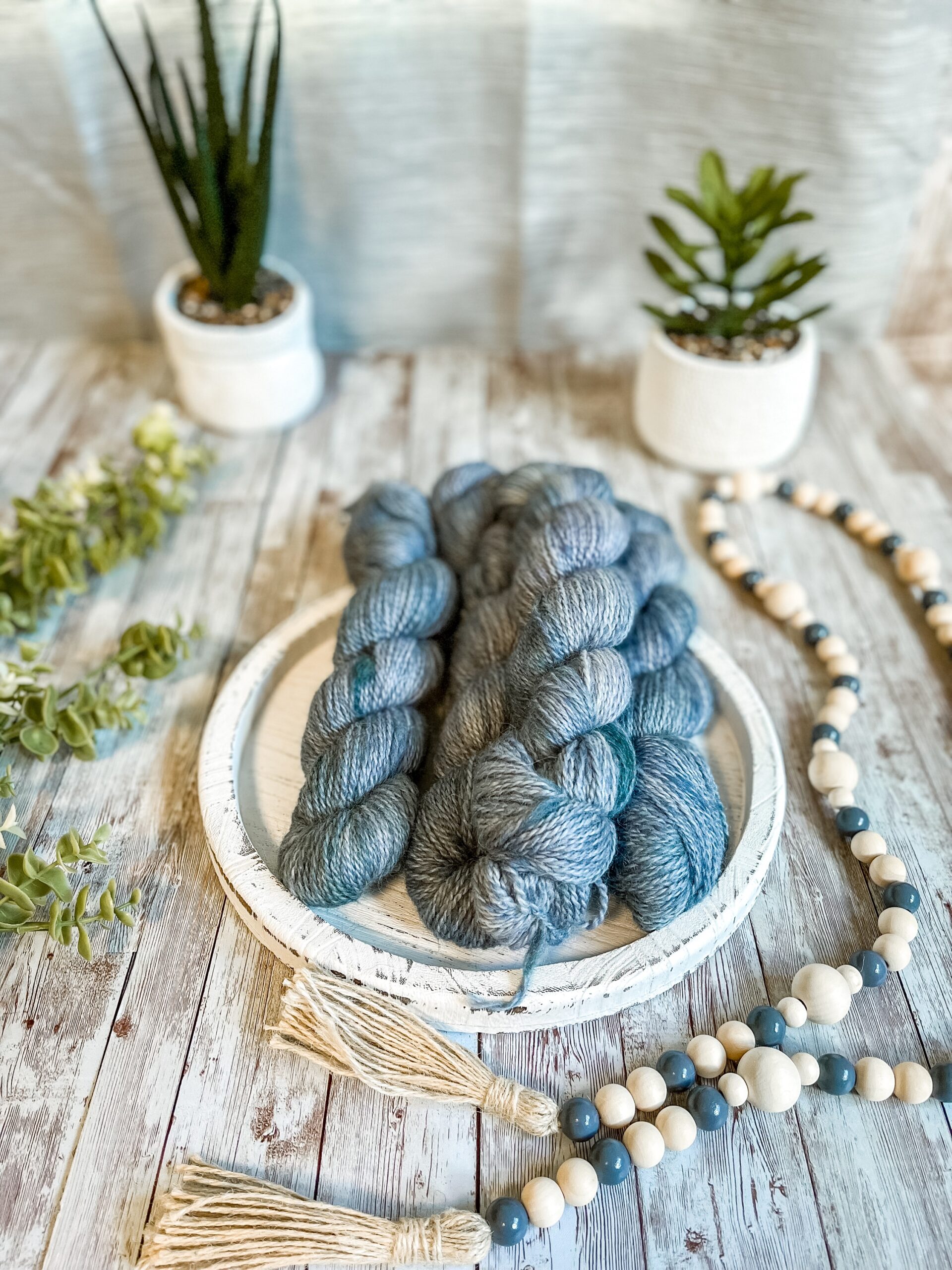 5 skeins of hand-dyed, blue yarn rest on a white wooden trivet. A strand of beads is draped to the right, some greenery is on the left, and 2 succulent plants are in the background.