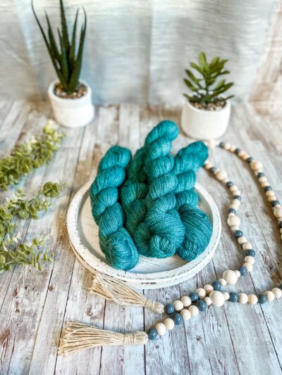 4 skeins of teal hand-dyed, yarn rest on a white wooden trivet. A strand of beads is draped to the right, some greenery is on the left, and 2 succulent plants are in the background.