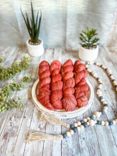 4 skeins of hand-dyed, watermelon red yarn rest on a white wooden trivet. A strand of beads is draped to the right, some greenery is on the left, and 2 succulent plants are in the background.