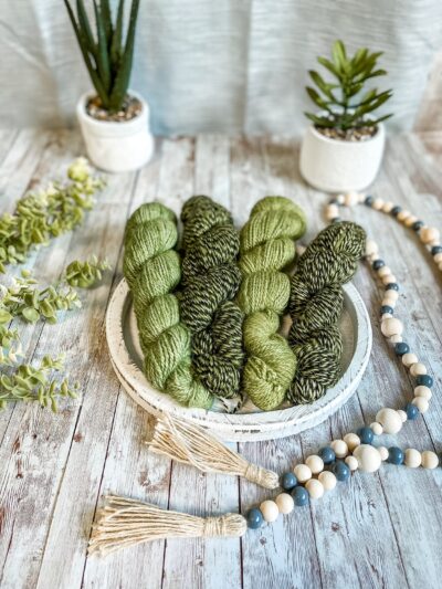 4 skeins of hand-dyed, moss green yarn and twisted moss green & black yarn rest on a white wooden trivet. A strand of beads is draped to the right, some greenery is on the left, and 2 succulent plants are in the background.