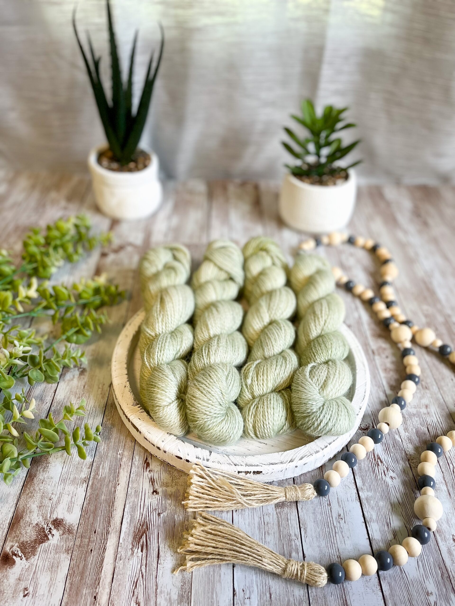 4 skeins of hand-dyed, pistachio green yarn rest on a white wooden trivet. A strand of beads is draped to the right, some greenery is on the left, and 2 succulent plants are in the background.