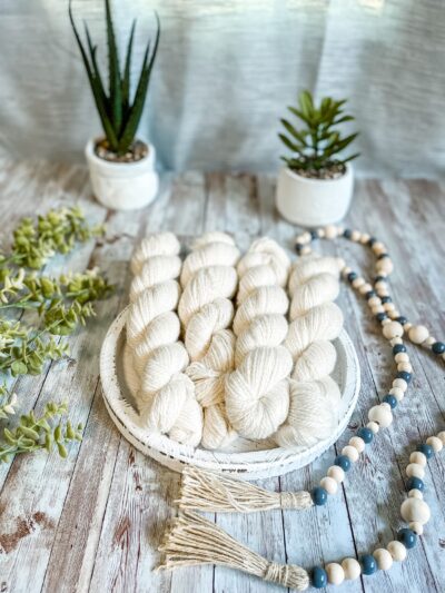 4 skeins of natural white yarn rest on a white wooden trivet. A strand of beads is draped to the right, some greenery is on the left, and 2 succulent plants are in the background.