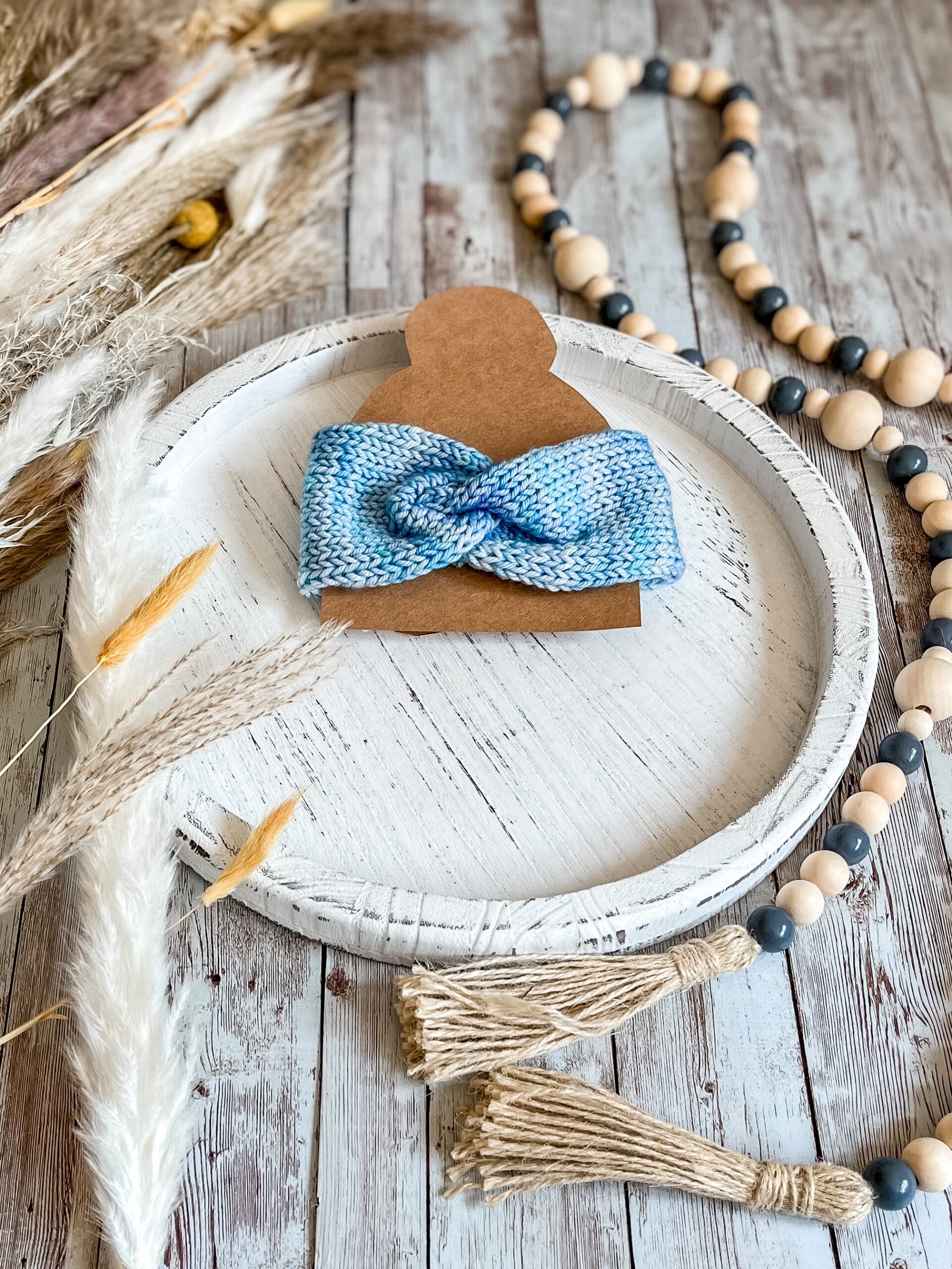 A blue, hand-dyed merino ear warmer is displayed on a kraft paper cutout of a messy bun on a whitewashed round wood platter. It is surrounded by wood beads on the right and faded pampas grass on the left, all on a wood plank panel