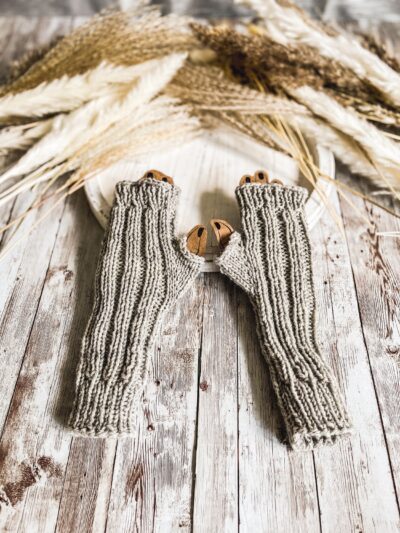 A pair of gray wool ribbed fingerless mitts is displayed on a rustic white wood trivet, with fall pampas grass stems in the backgound