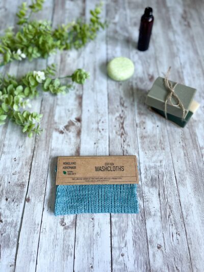 A blue Virginia grown cotton washcloth is displayed with a tag while resting on a wood plank, surrounded by eucalyptus greenery, a set of 2 bar soaps, a round shampoo bar and an amber bottle of wash