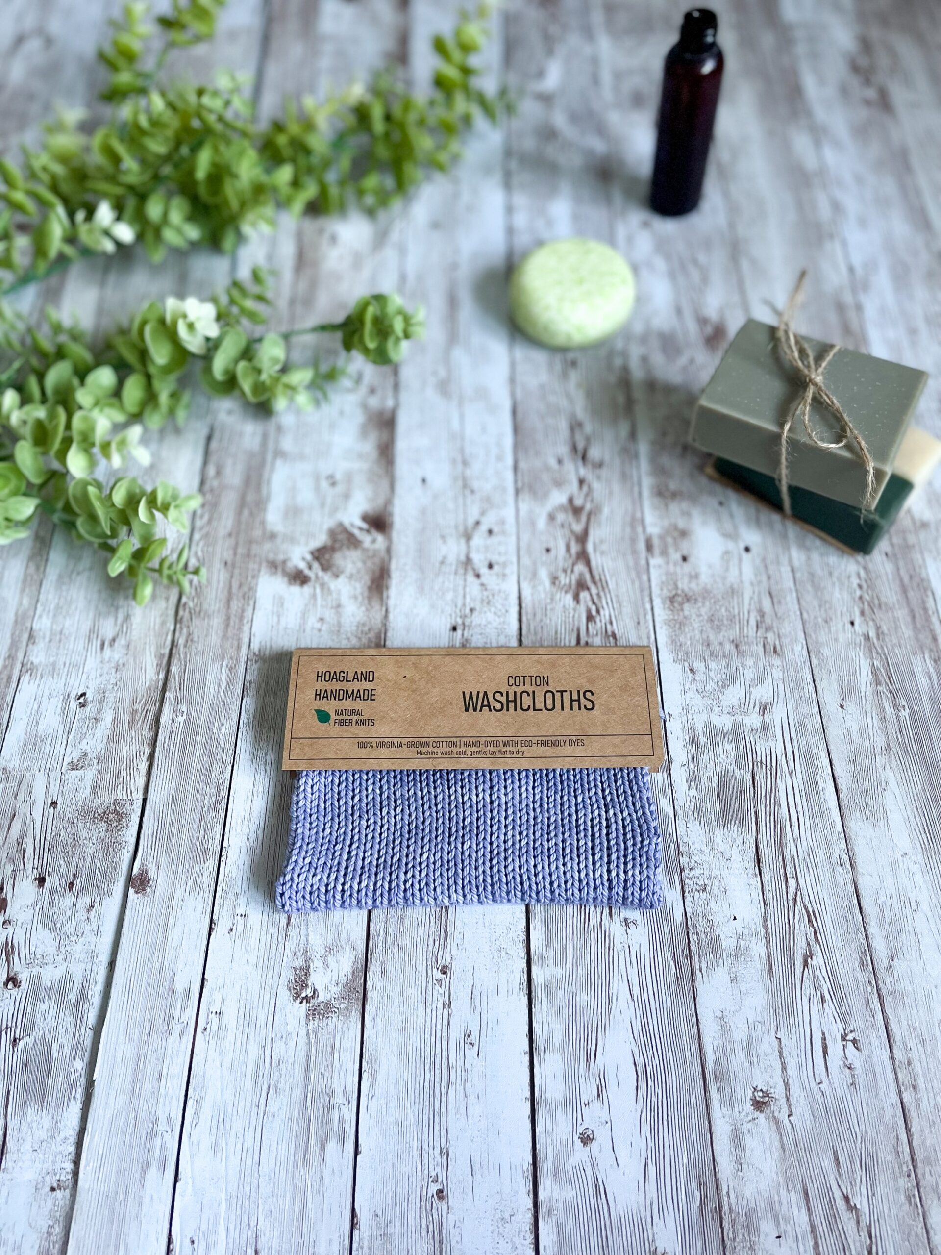 A purple Virginia grown cotton washcloth is displayed with a tag while resting on a wood plank, surrounded by eucalyptus greenery, a set of 2 bar soaps, a round shampoo bar and an amber bottle of wash
