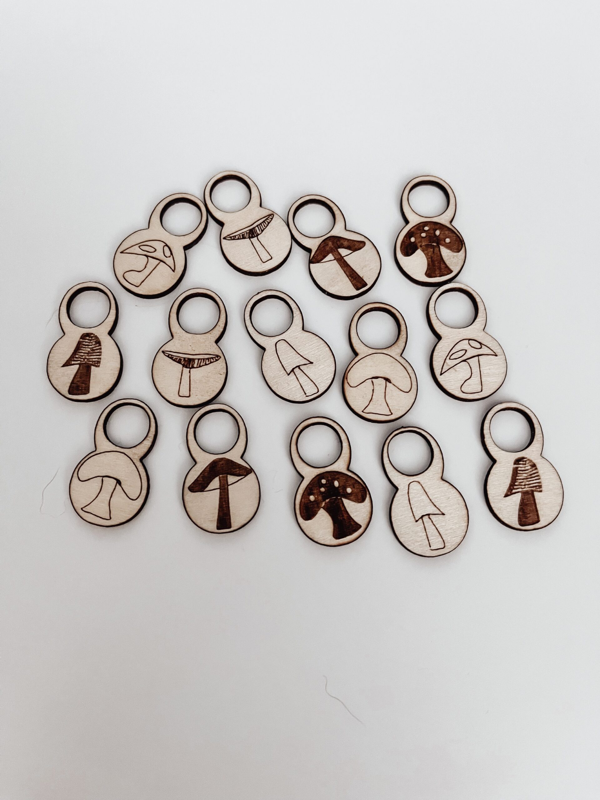 A total of 14 wood stitch markers with a variety of hand-drawn mushroom designs are displayed on a white background.