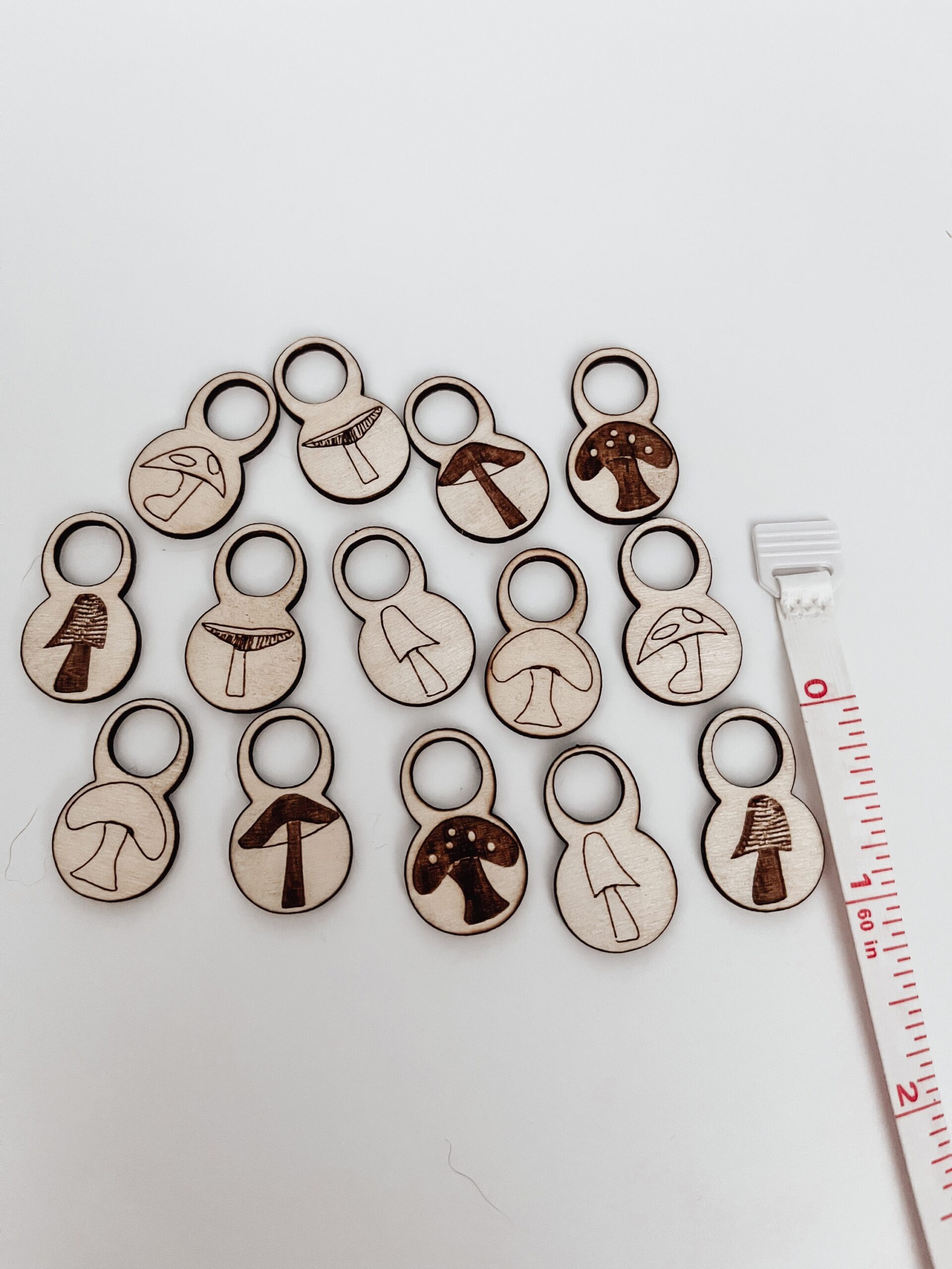 A total of 14 wood stitch markers with a variety of hand-drawn mushroom designs are displayed on a white background. next to a measuring tape showing they're approximately 1" tall.
