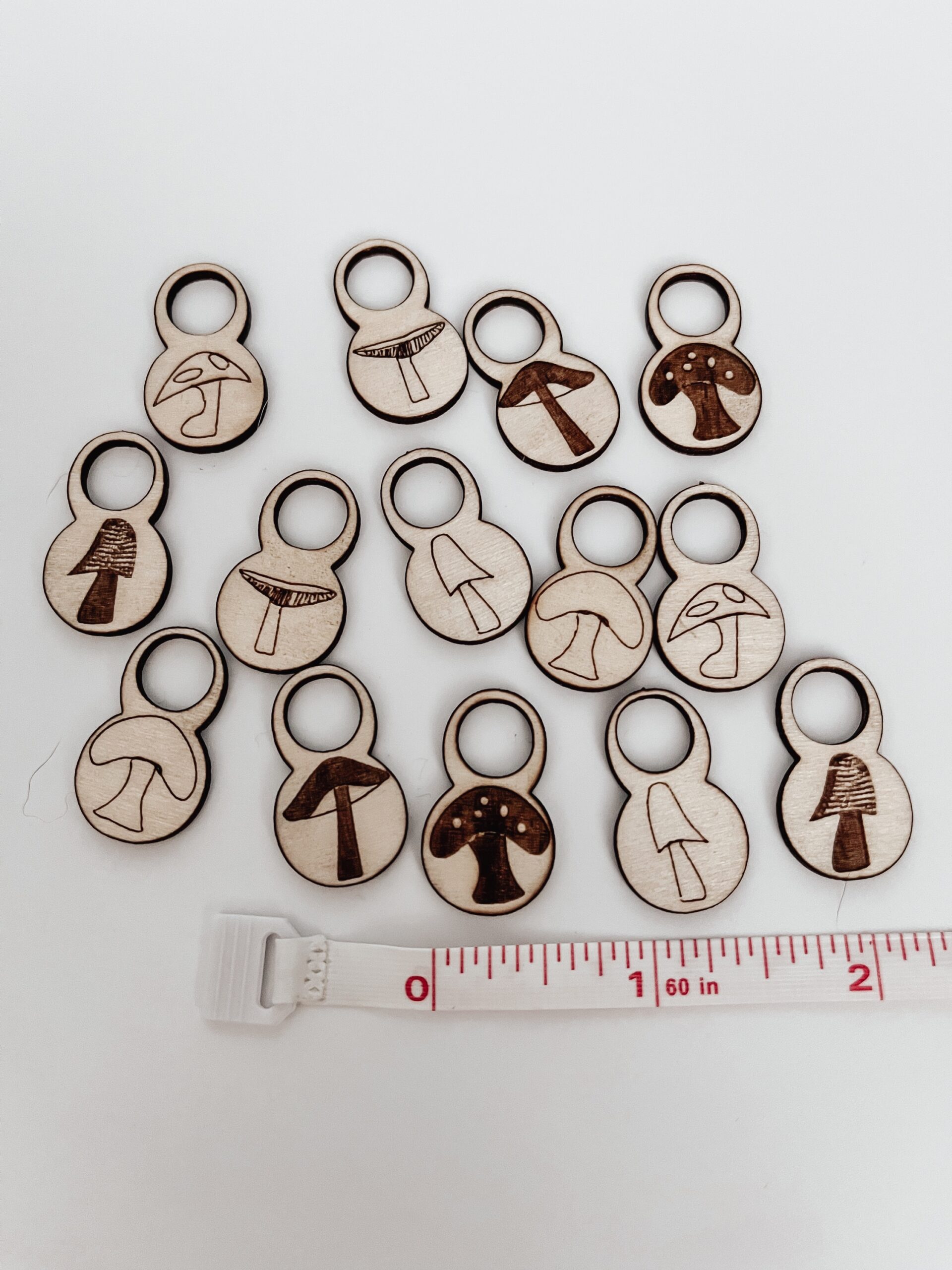 A total of 14 wood stitch markers with a variety of hand-drawn mushroom designs are displayed on a white background. next to a measuring tape showing they're approximately 1/2" wide