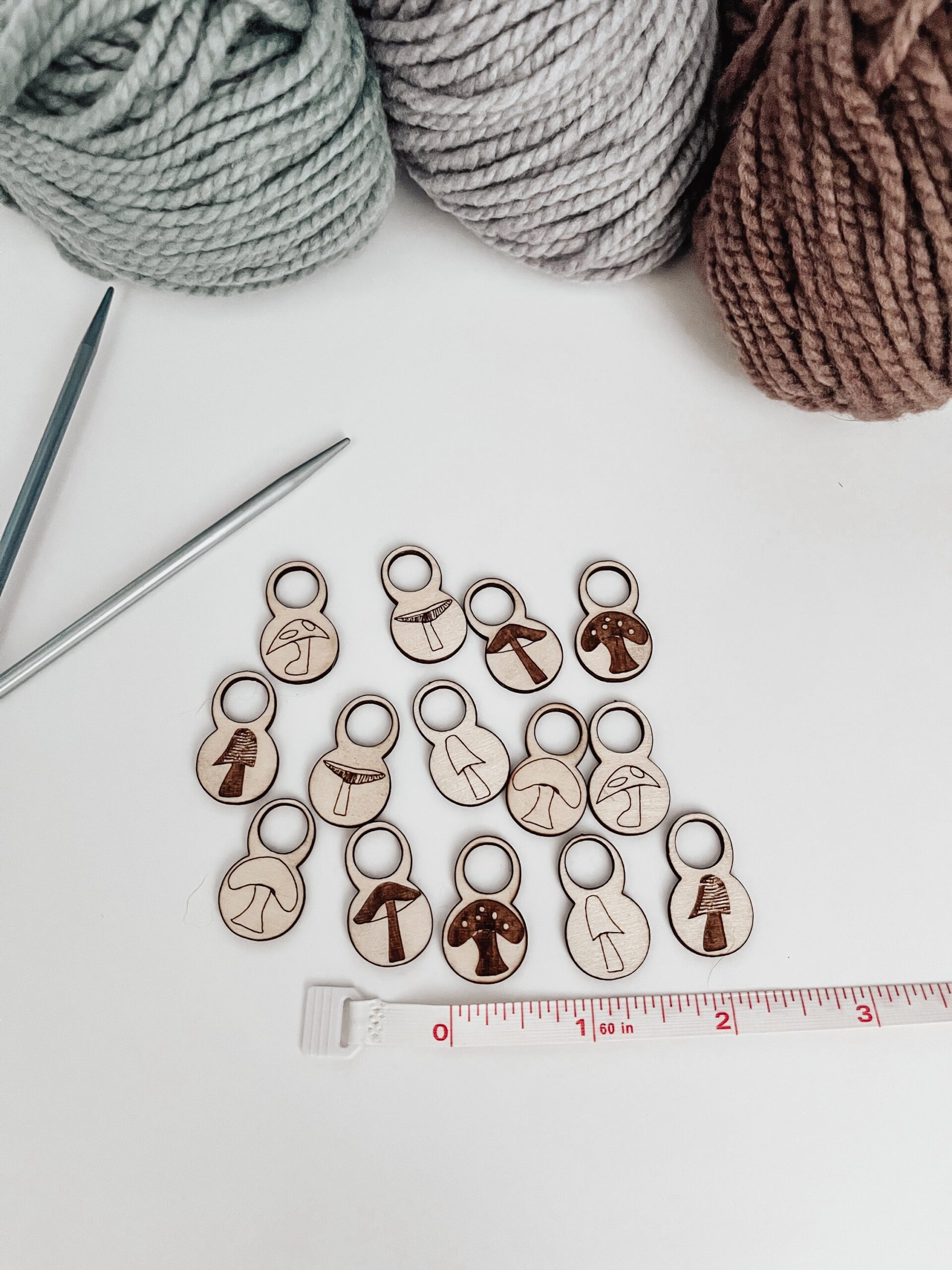 A total of 14 wood stitch markers with a variety of hand-drawn mushroom designs are displayed next to a measuring tape that shows they're approximately 1/4" wide. A pair of metal knitting needles and three skeins of merino yarn are in the background.