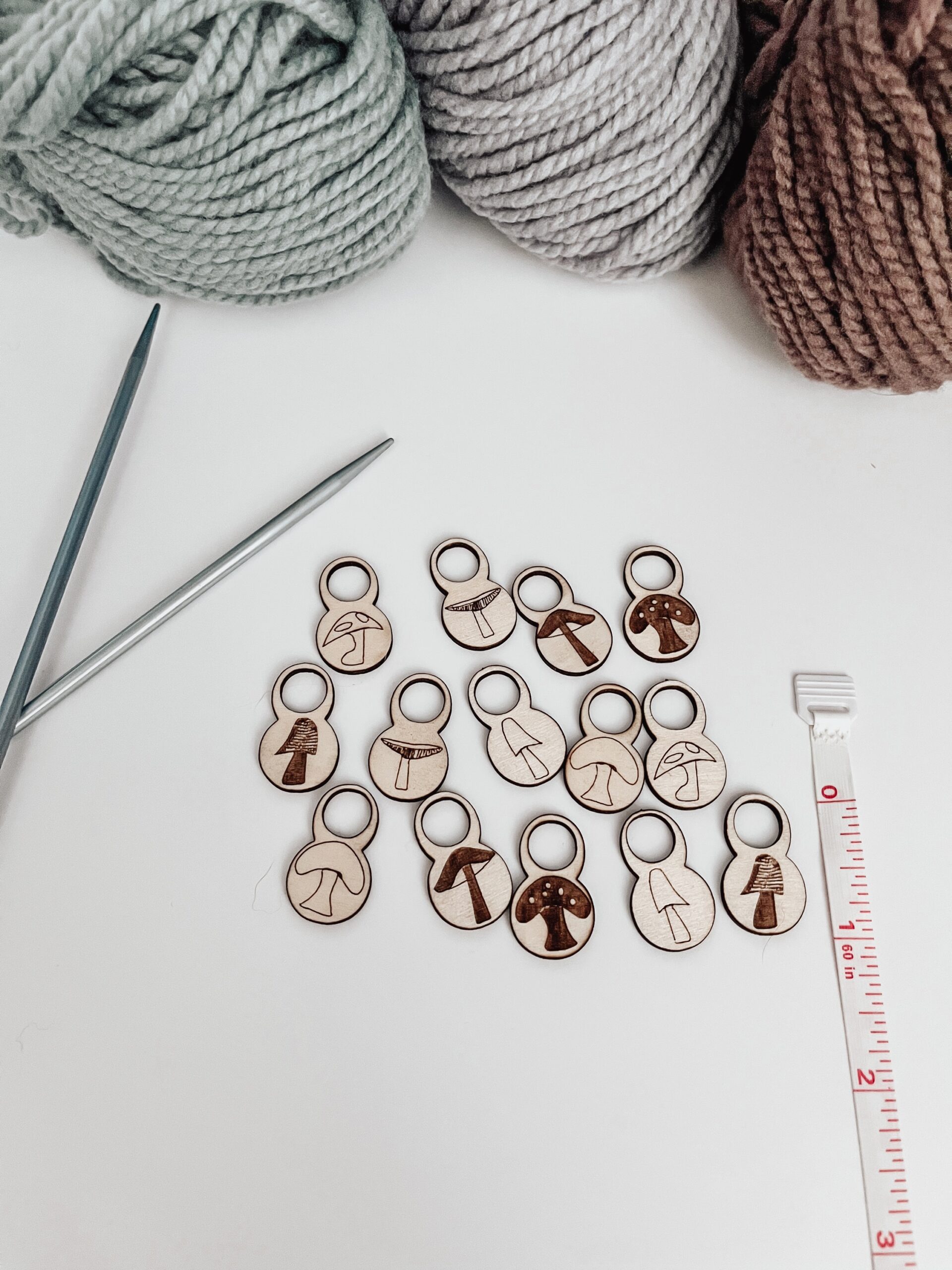 A total of 14 wood stitch markers with a variety of hand-drawn mushroom designs are displayed next to a measuring tape that shows they're approximately 1/2" tall. A pair of metal knitting needles and three skeins of merino yarn are in the background.